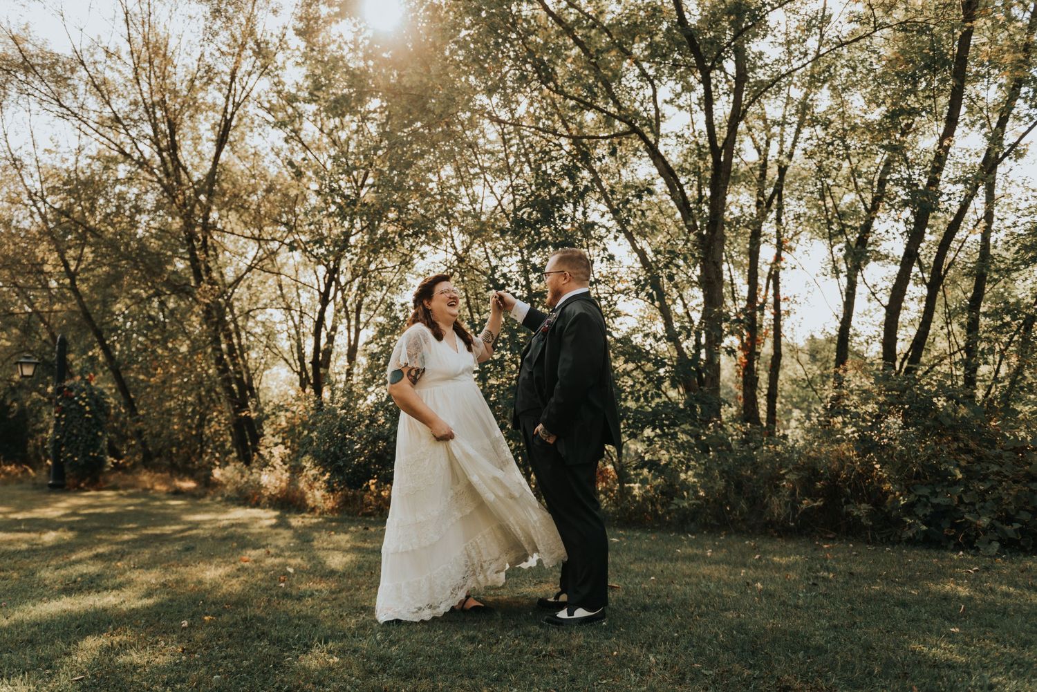 A couple in formal wedding attire stands in a sunlit wooded area on a beautiful autumn afternoon.