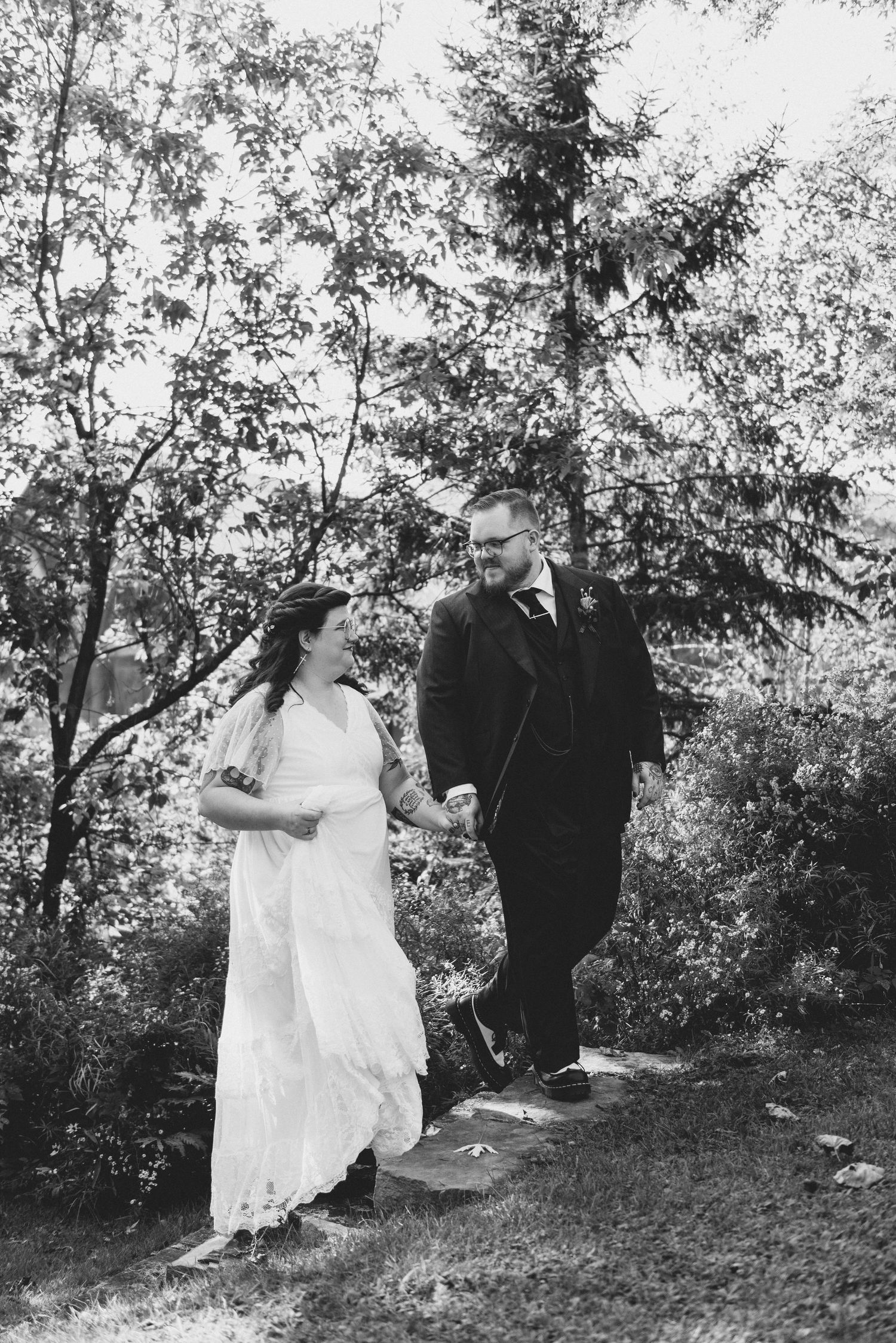 A black and white photograph of a couple walking together on a wooded path, capturing an outdoor wedding moment.