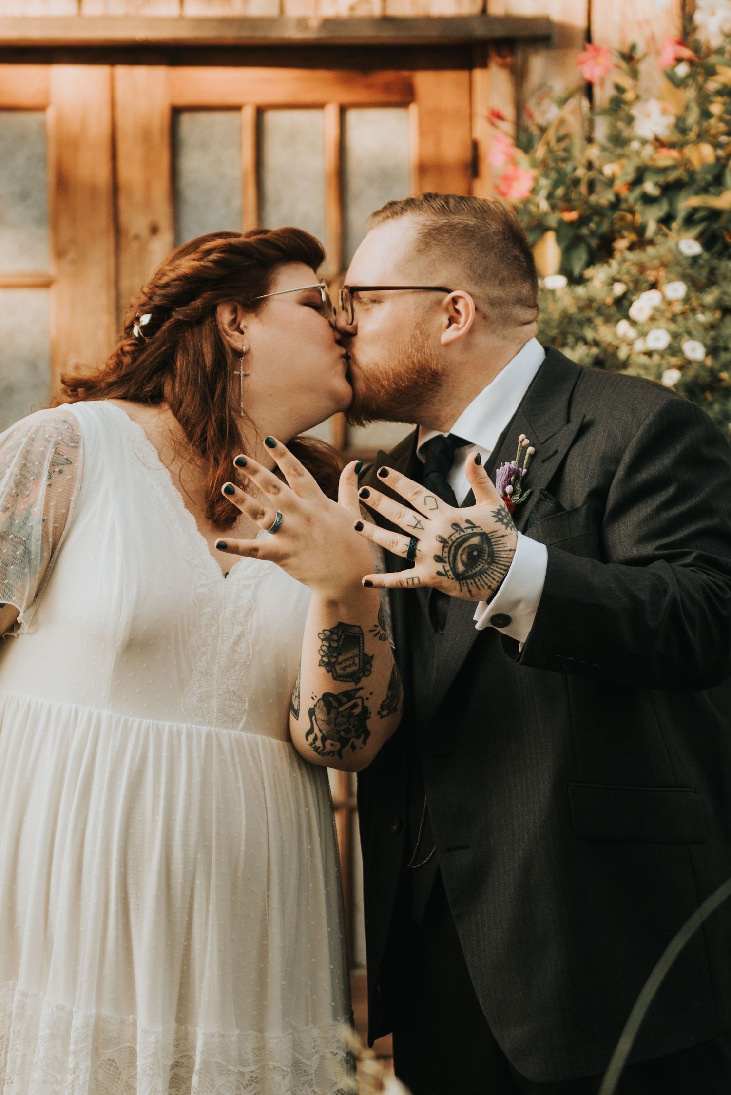 A couple shares a tender kiss at their wedding, showcasing their tattooed hands and wedding attire in warm lighting.