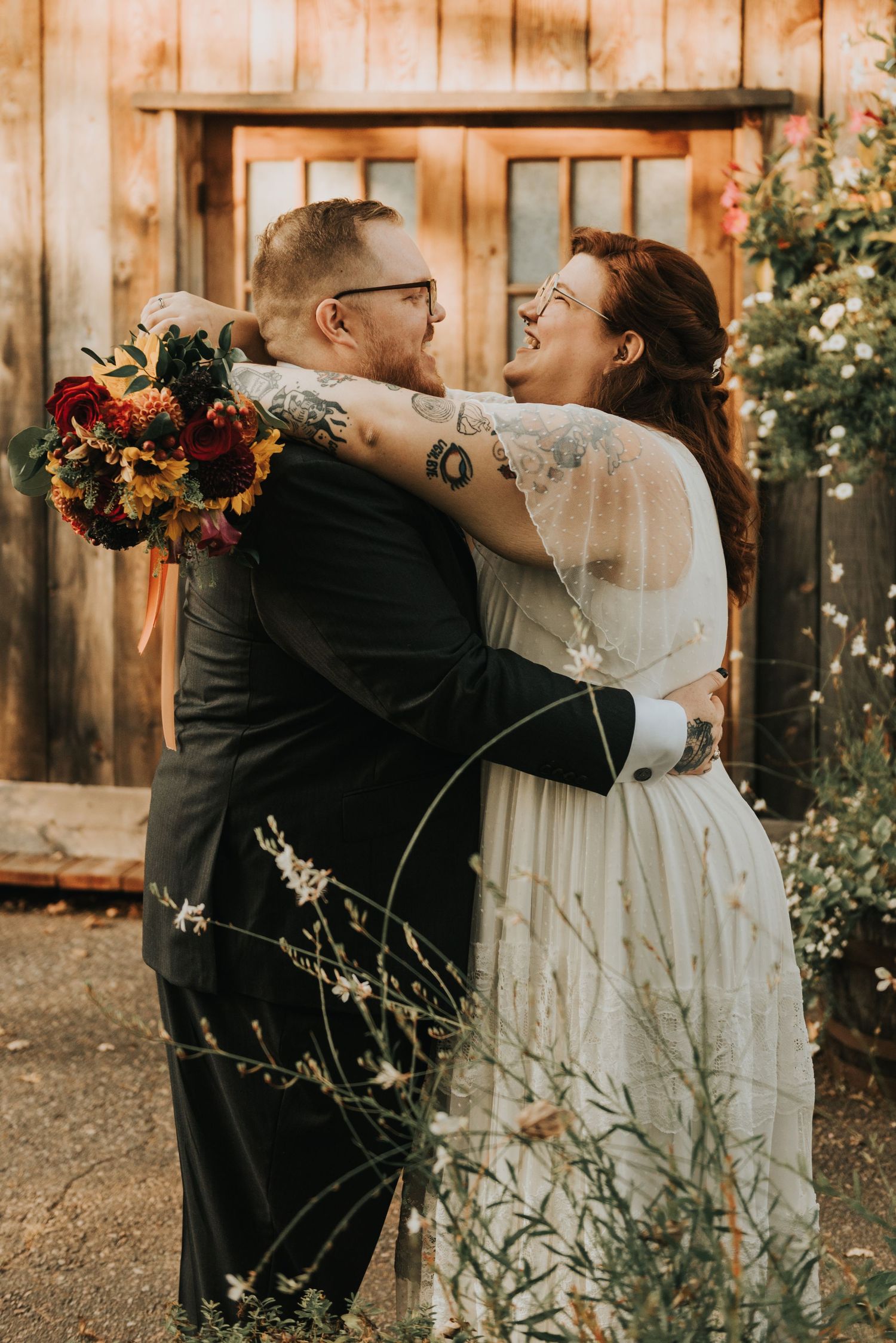Intimate outdoor wedding portrait with autumn bouquet against rustic wooden barn backdrop in warm golden light.