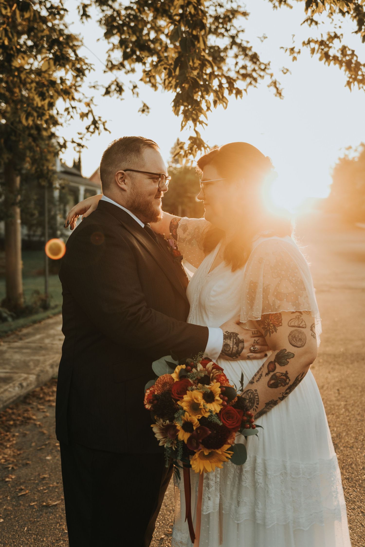 A romantic wedding photo of a couple embracing at sunset with a sunflower bouquet and autumn foliage overhead.