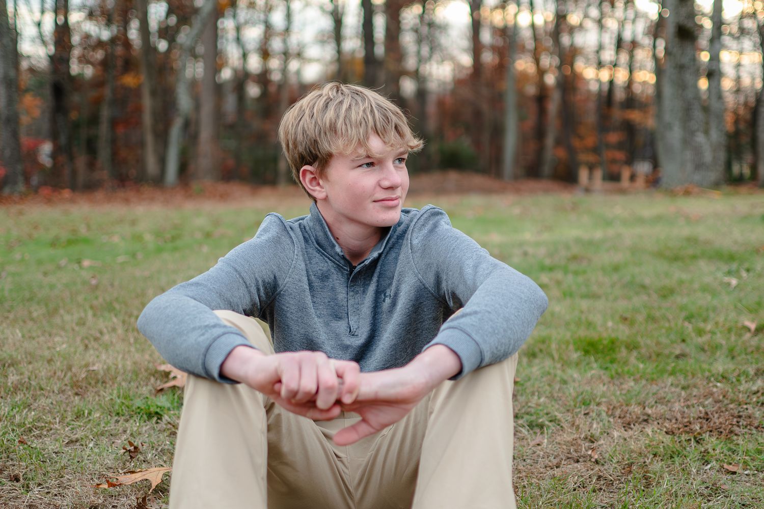 A person in a gray sweater sits on the ground in an autumnal outdoor setting with trees in the background.