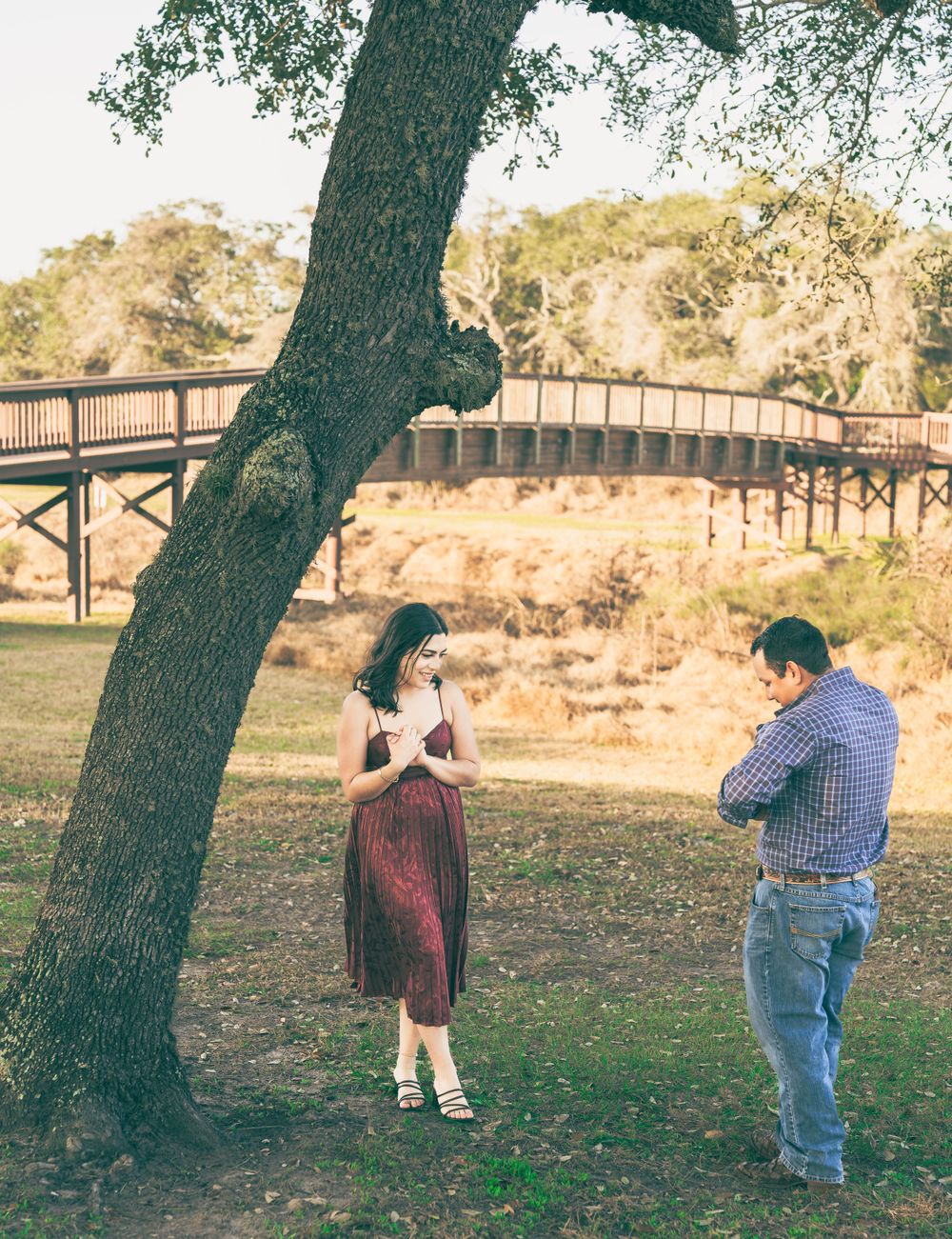A couple poses under a large tree near a rustic bridge in a natural park setting during golden hour.
