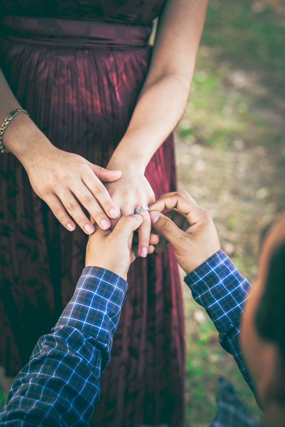 A close-up shot of hands placing an engagement ring on a finger, with a burgundy dress in the background.
