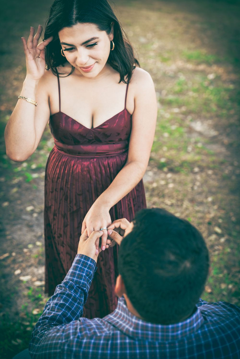 An intimate proposal shot with person in burgundy dress reacting as someone offers ring outdoors.