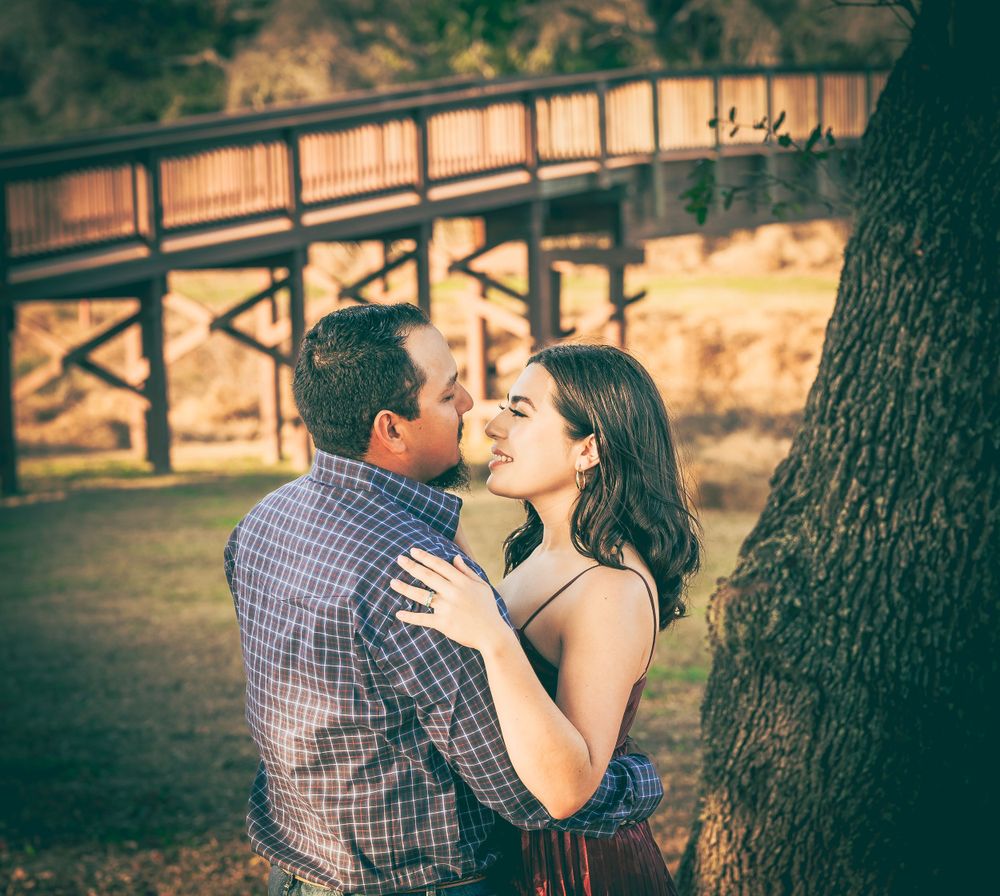 A romantic couple embrace near a wooden bridge in a park during golden hour.