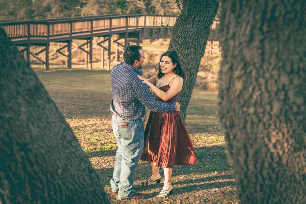 A couple dances together under a large tree with a wooden bridge visible in the background.