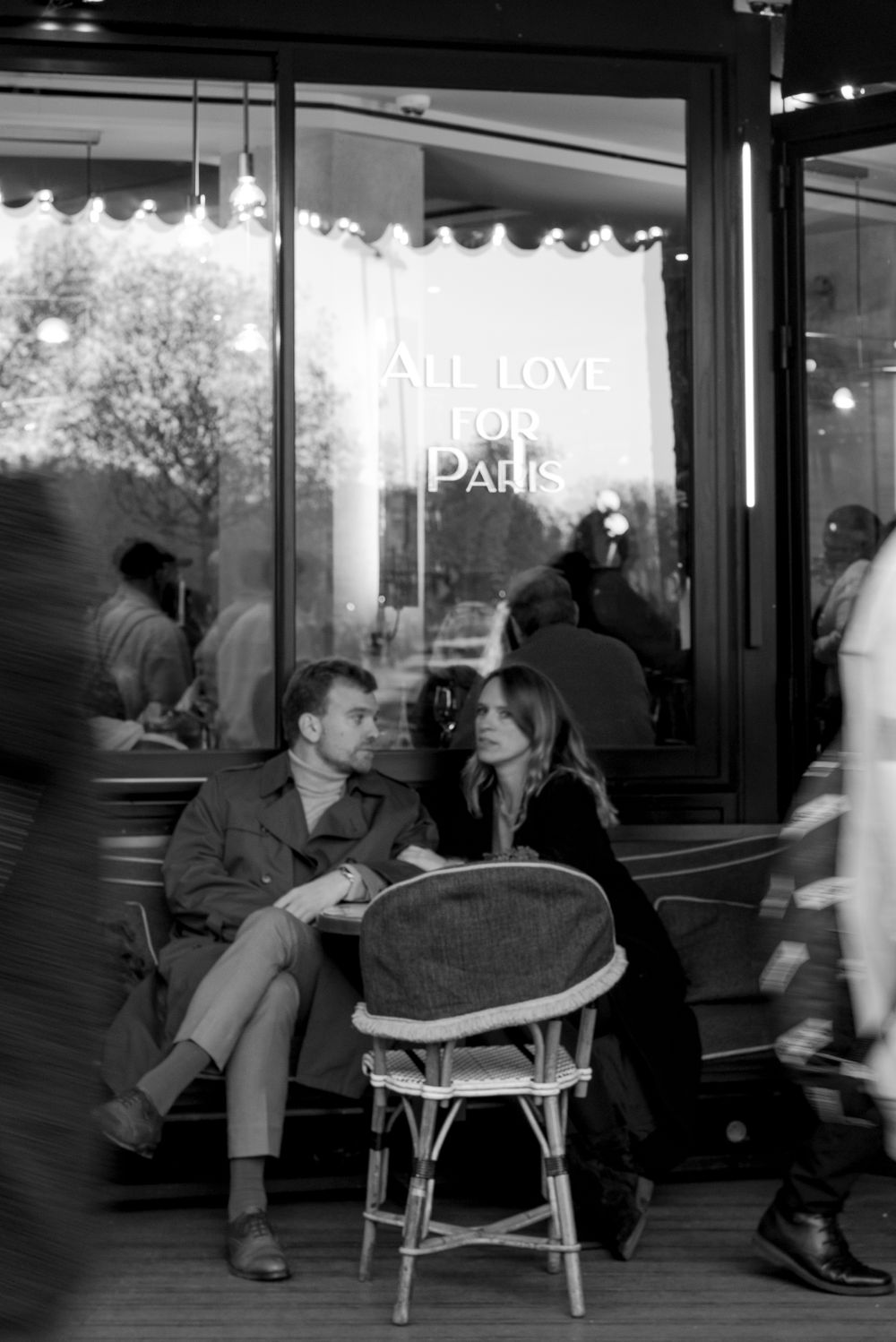 A black and white photo of a couple sitting at a Parisian cafe with a neon sign reading "ALL LOVE PARIS" in the window.