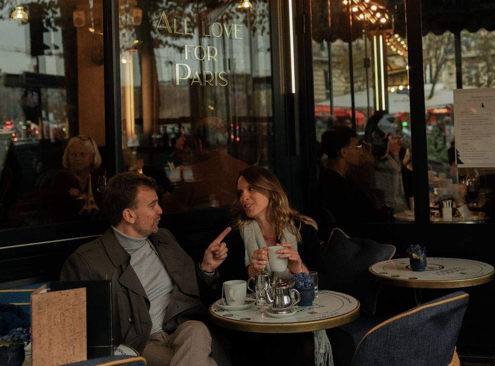 Two people engaged in conversation at a cozy Parisian cafe with outdoor seating and city reflections in the window.