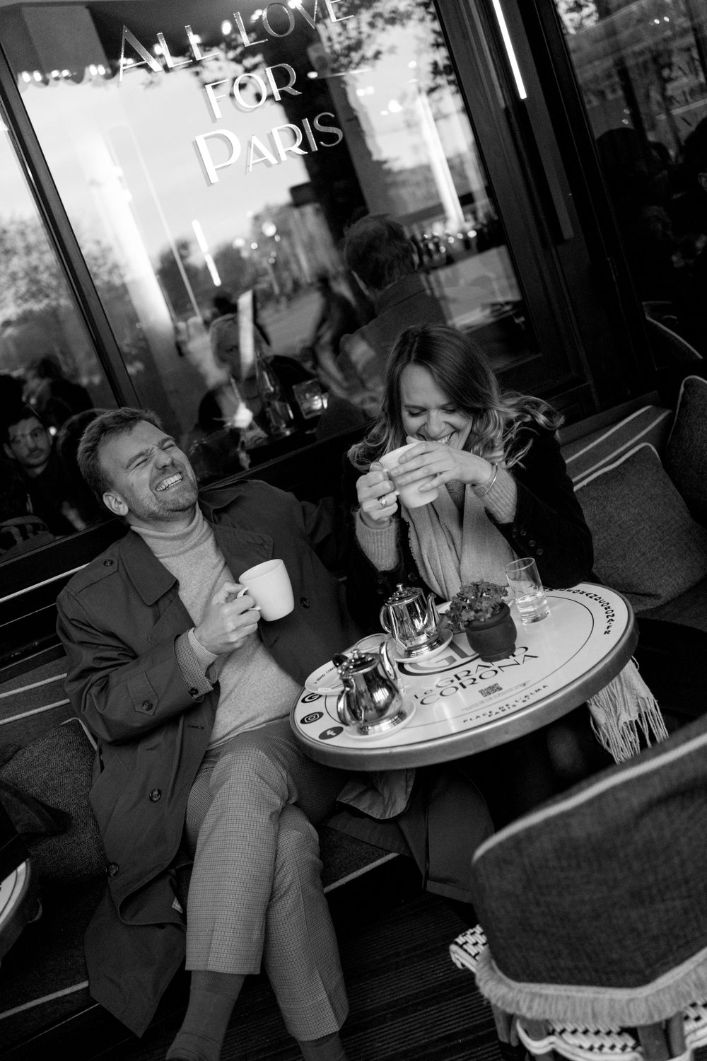 Two people share a lighthearted moment while enjoying drinks at a Paris cafe terrace in black and white photography.
