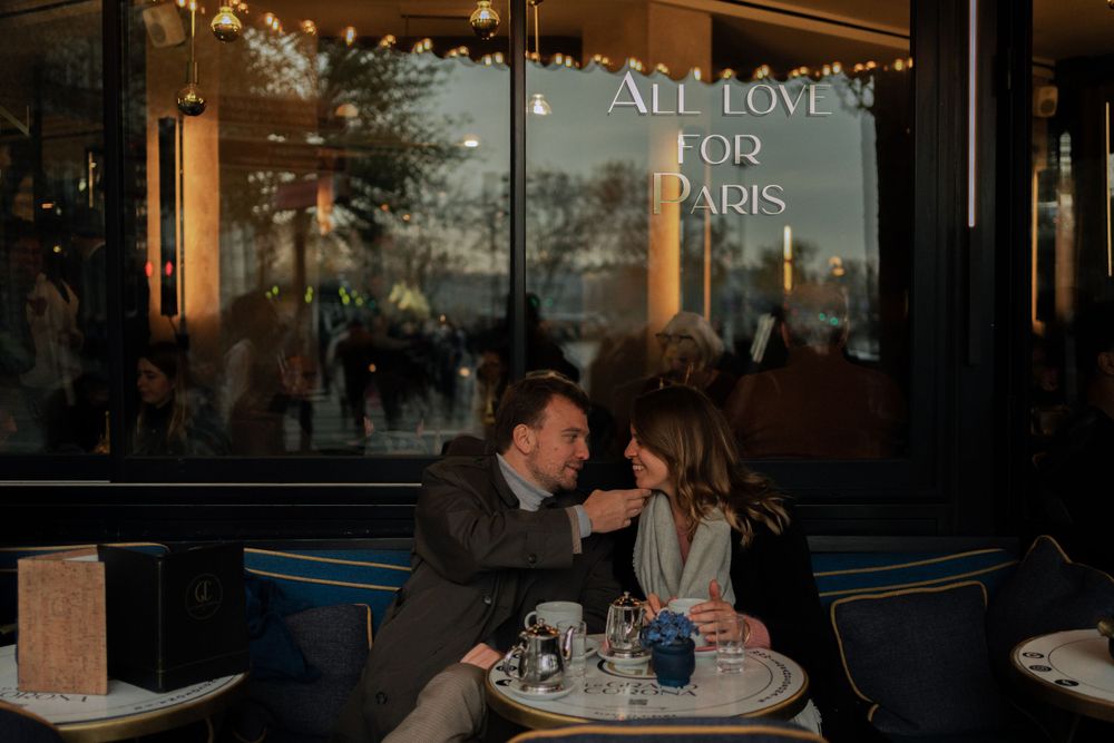A romantic couple shares an intimate moment at an outdoor cafe table with neon lights and storefront reflections in the background.