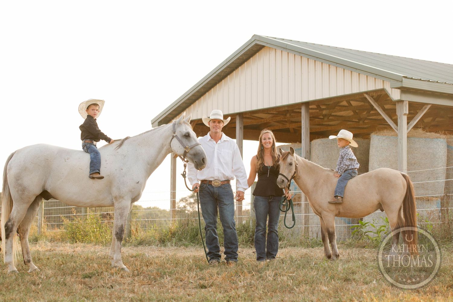 White horses and family members gather near a wooden barn at sunset.
