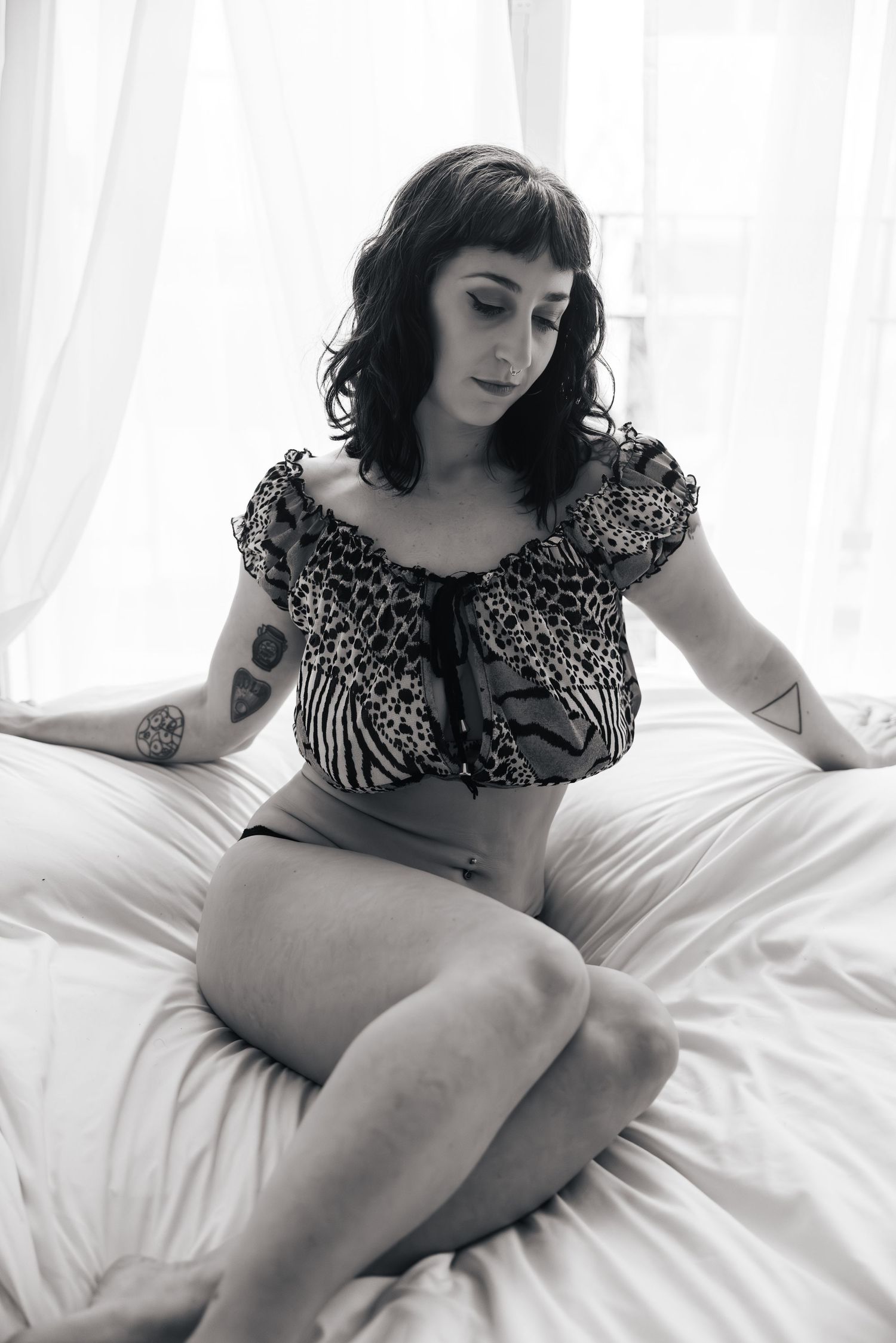 Black and white boudoir photo with woman in patterned crop top reclining on white bedding.