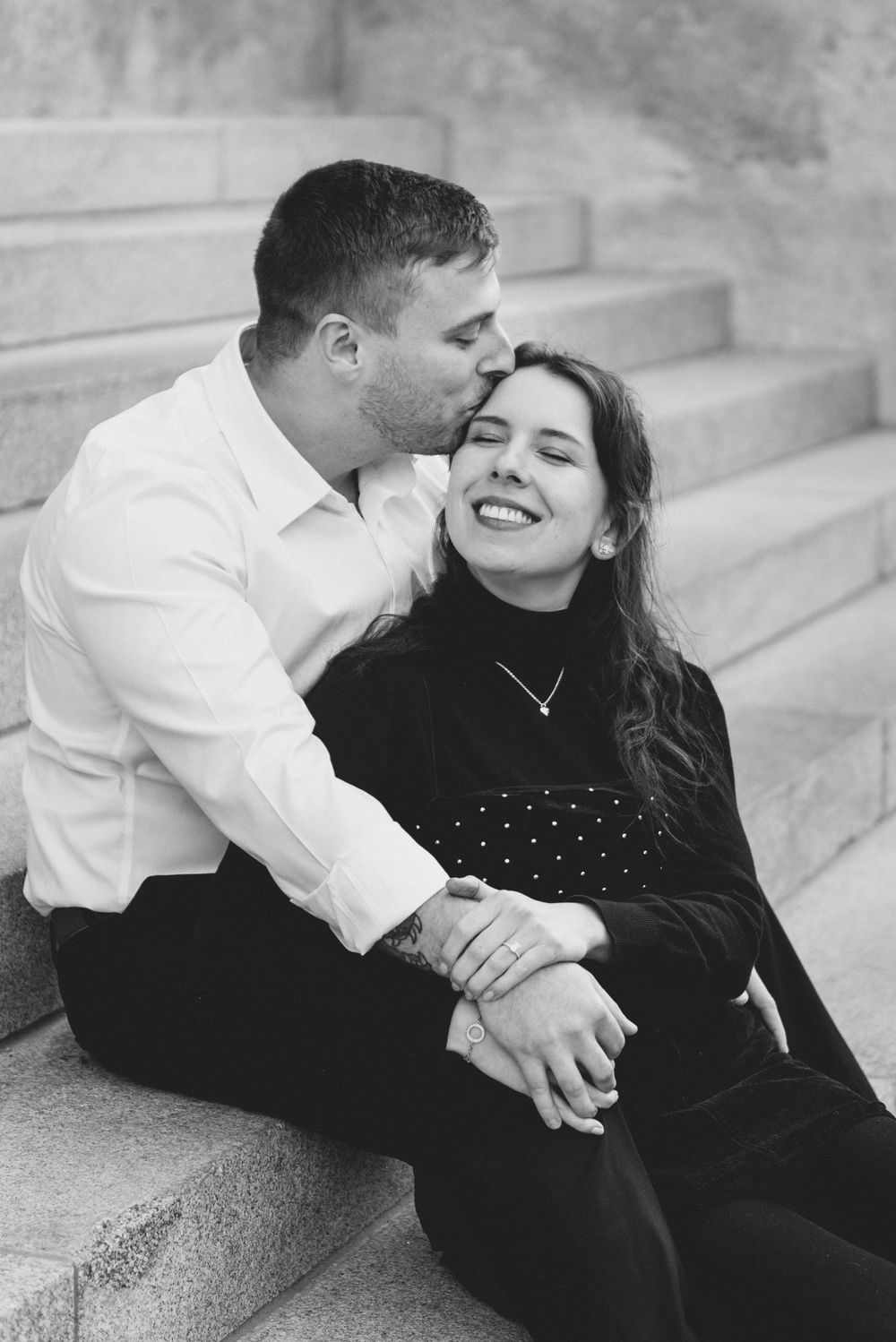 A romantic black and white photo of a couple sharing a joyful moment while sitting on concrete steps outdoors.