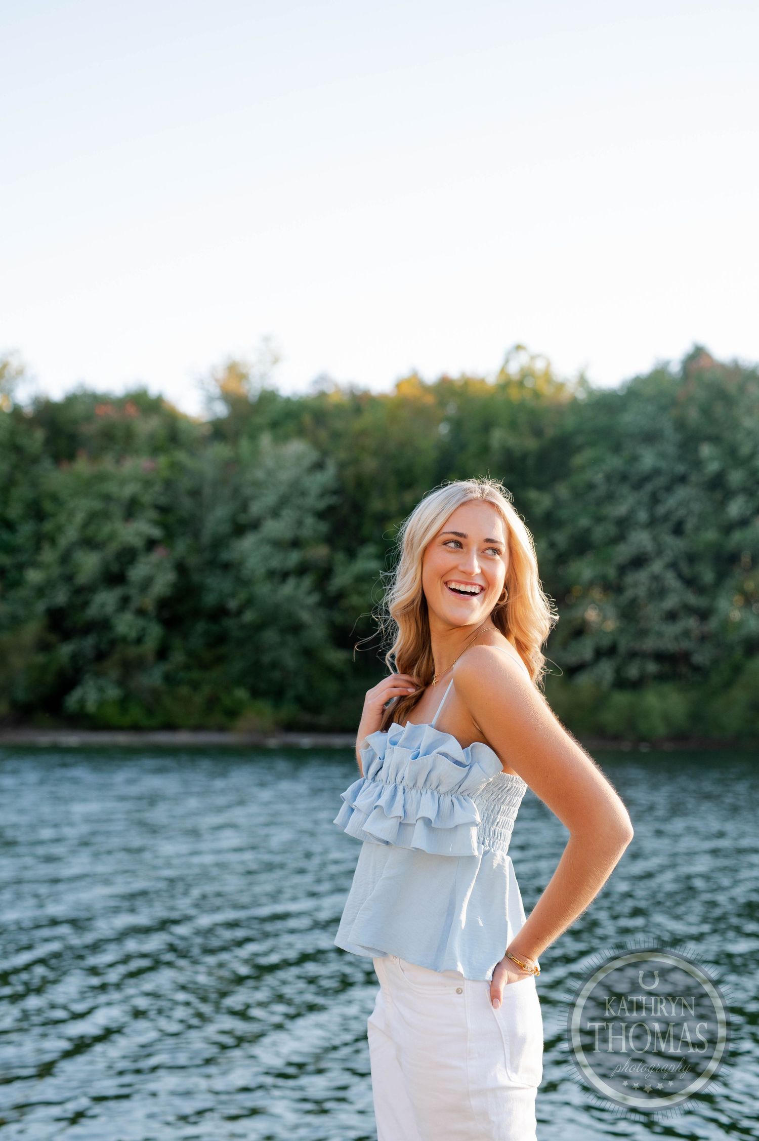 Joyful summer portrait of someone in a ruffled blue top standing on a dock by a lake at sunset.