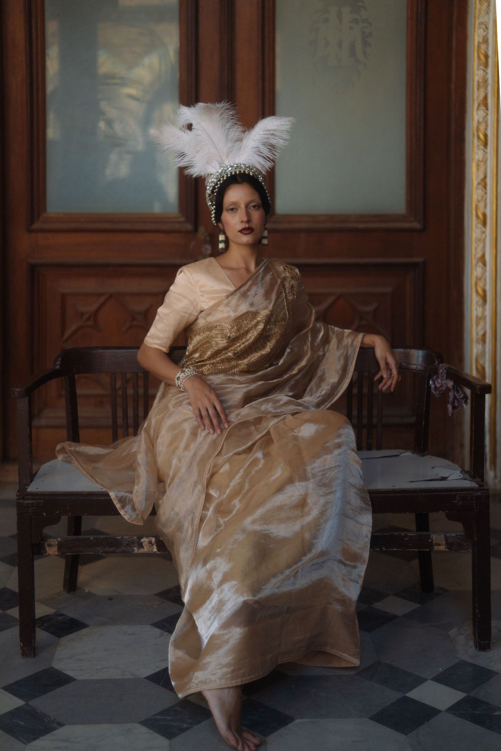 Elegant portrait in beige sari with white feather headpiece on checkered floor.