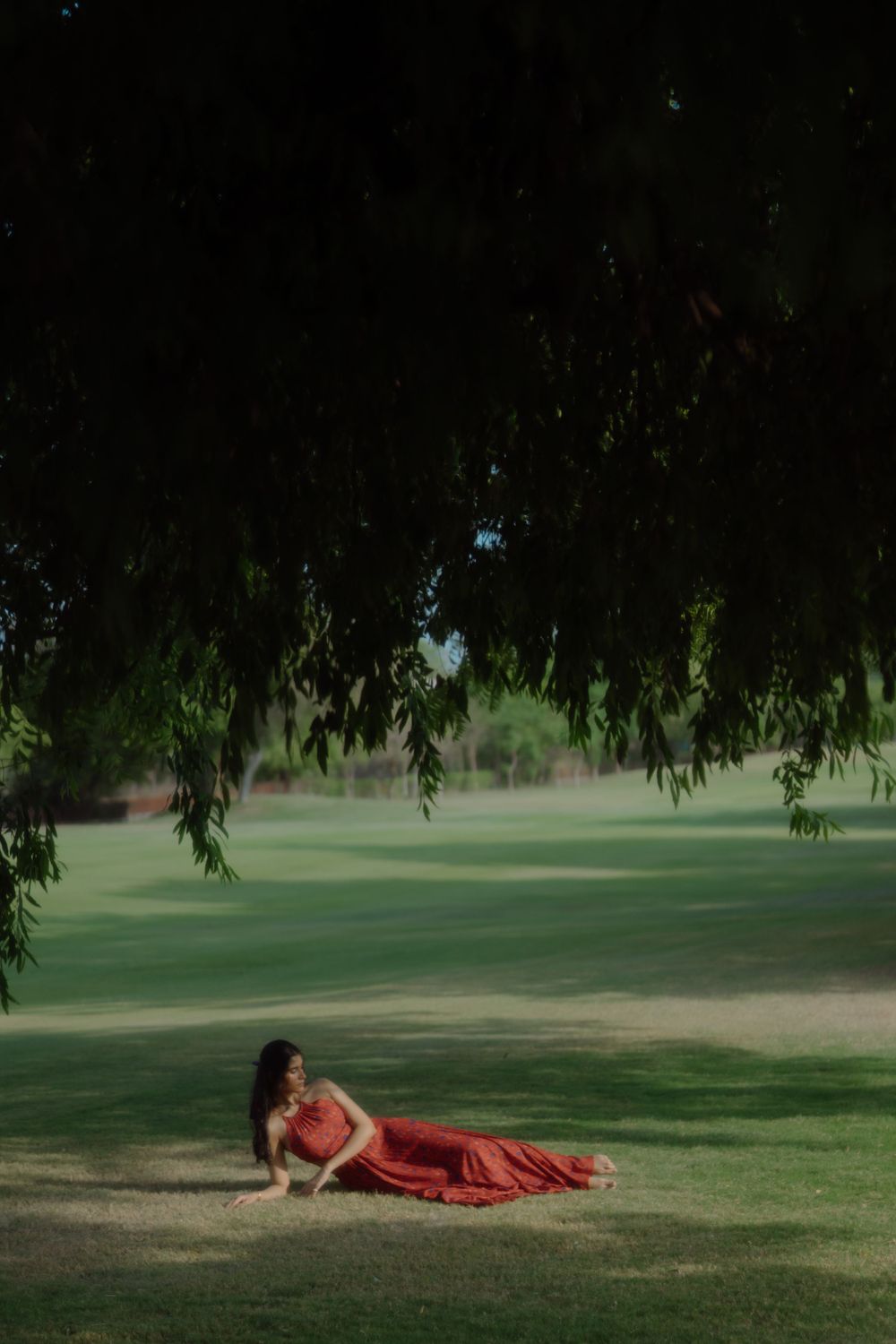 Figure in red dress reclining on grass under weeping willow branches.