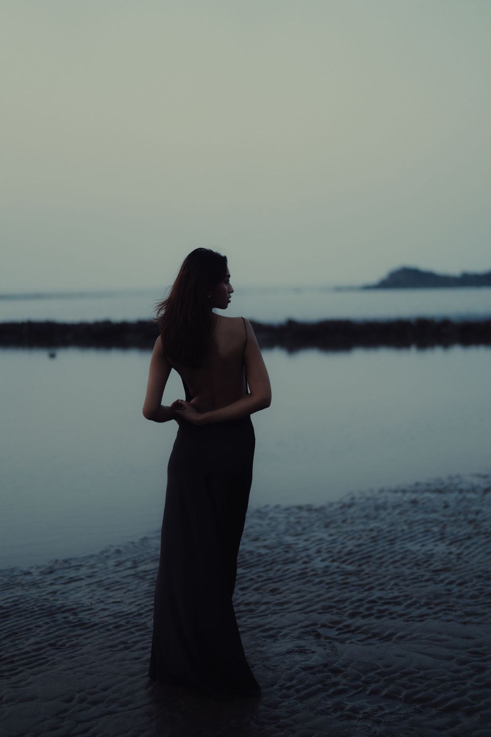 Silhouette of figure in long dress standing by calm water at dusk.
