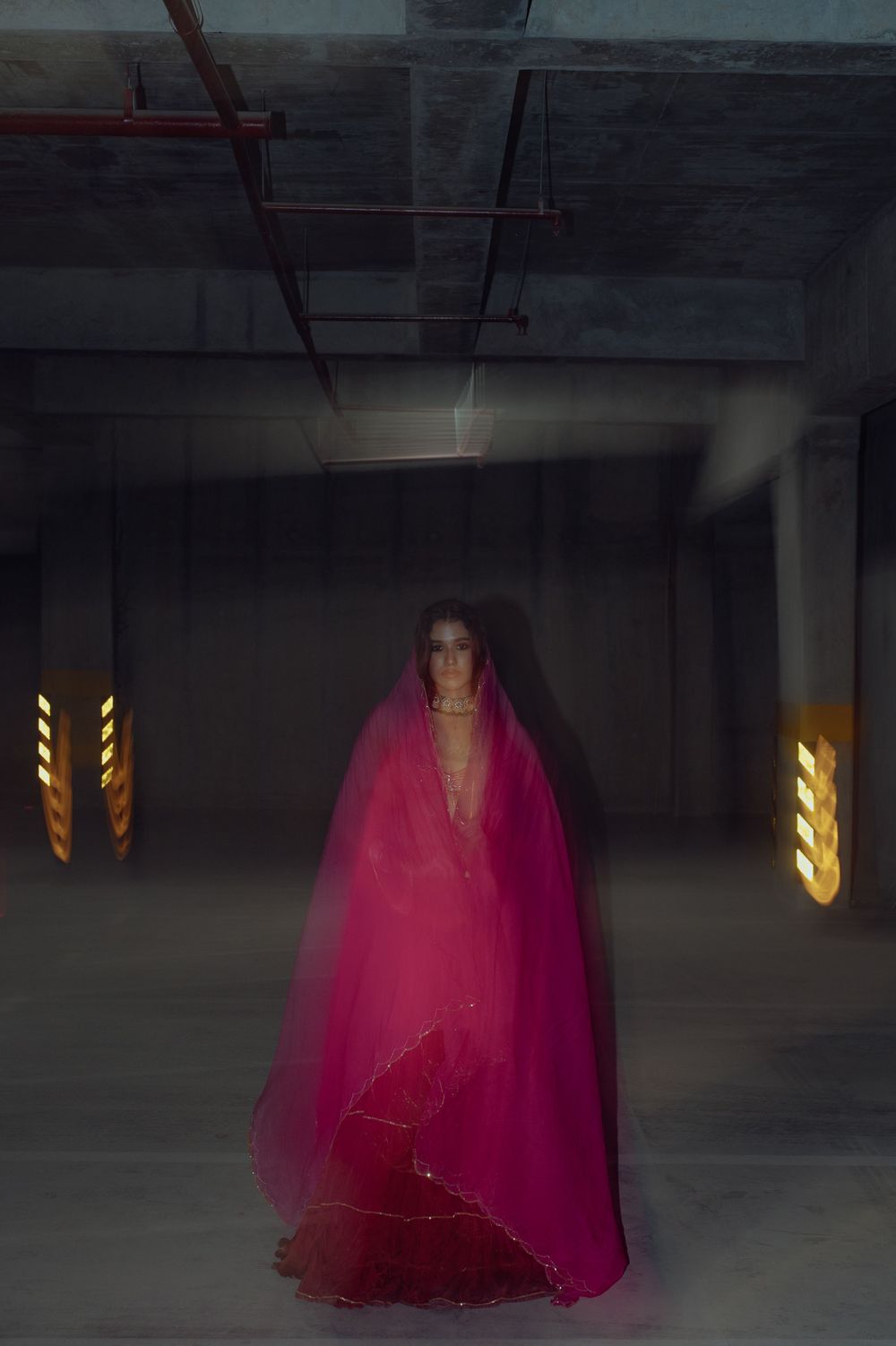 A dramatic figure in a flowing bright pink gown stands in a dimly lit parking garage with yellow lights.