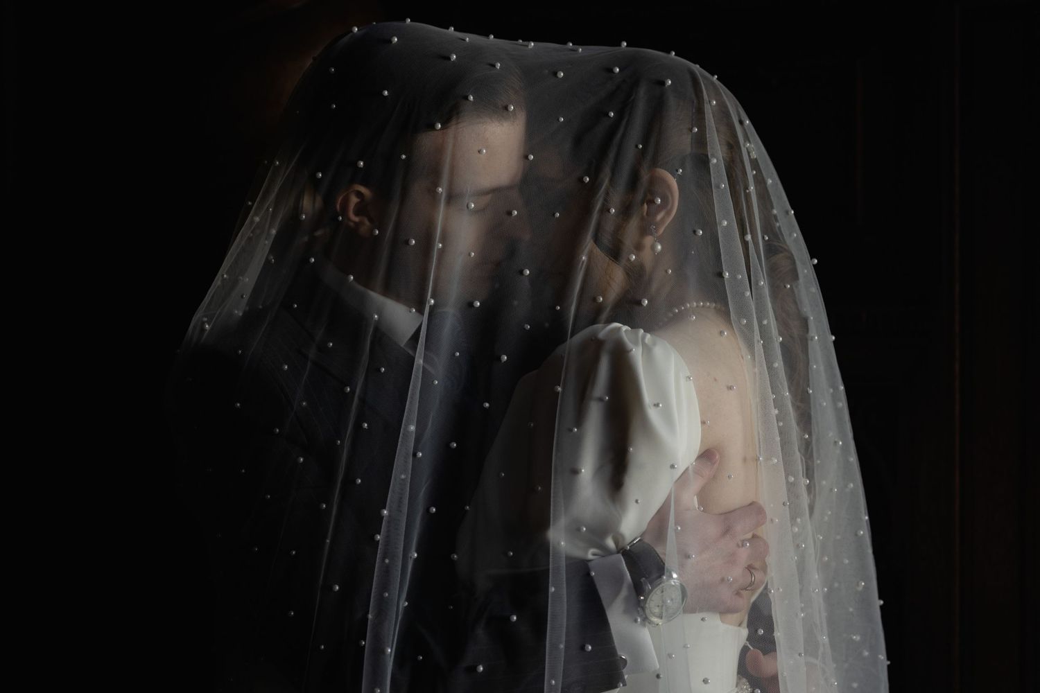 A romantic side view of a couple embracing under a white veil against a dark background.