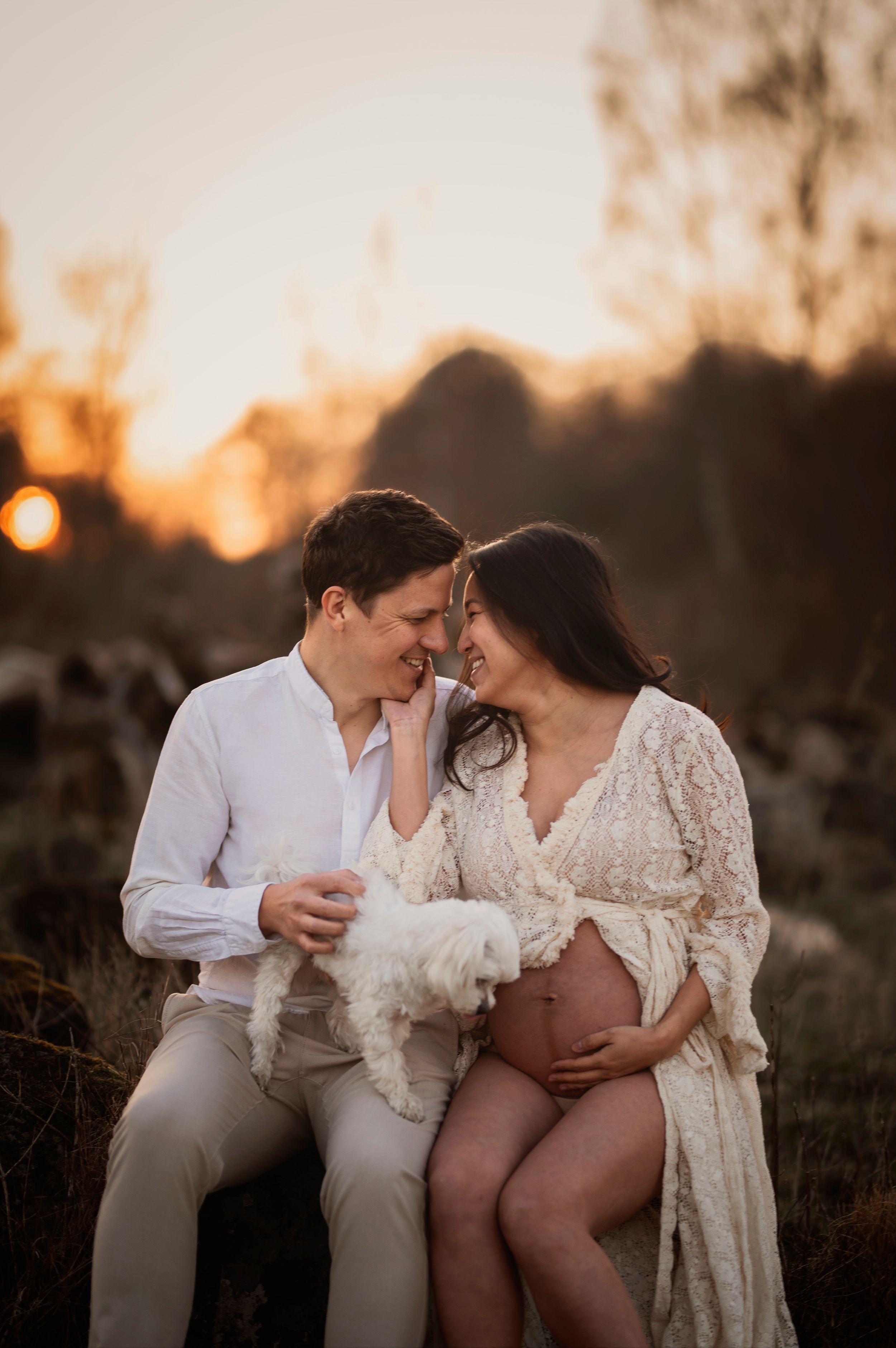 Romantic maternity photo shoot at sunset with couple and small white dog sharing intimate moment in nature.