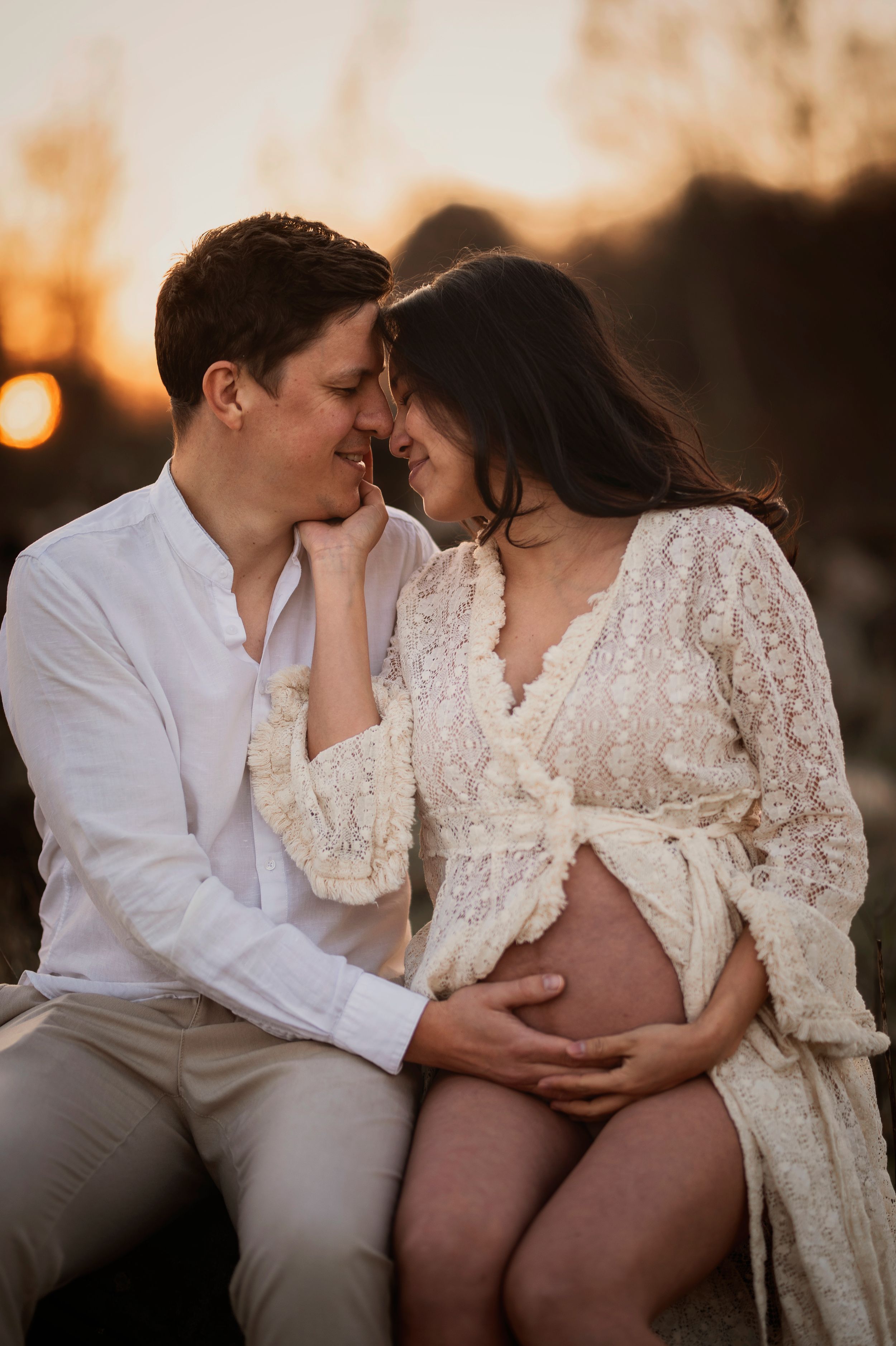 A tender maternity photo of a couple sharing an intimate moment at sunset in white lace and formal attire.
