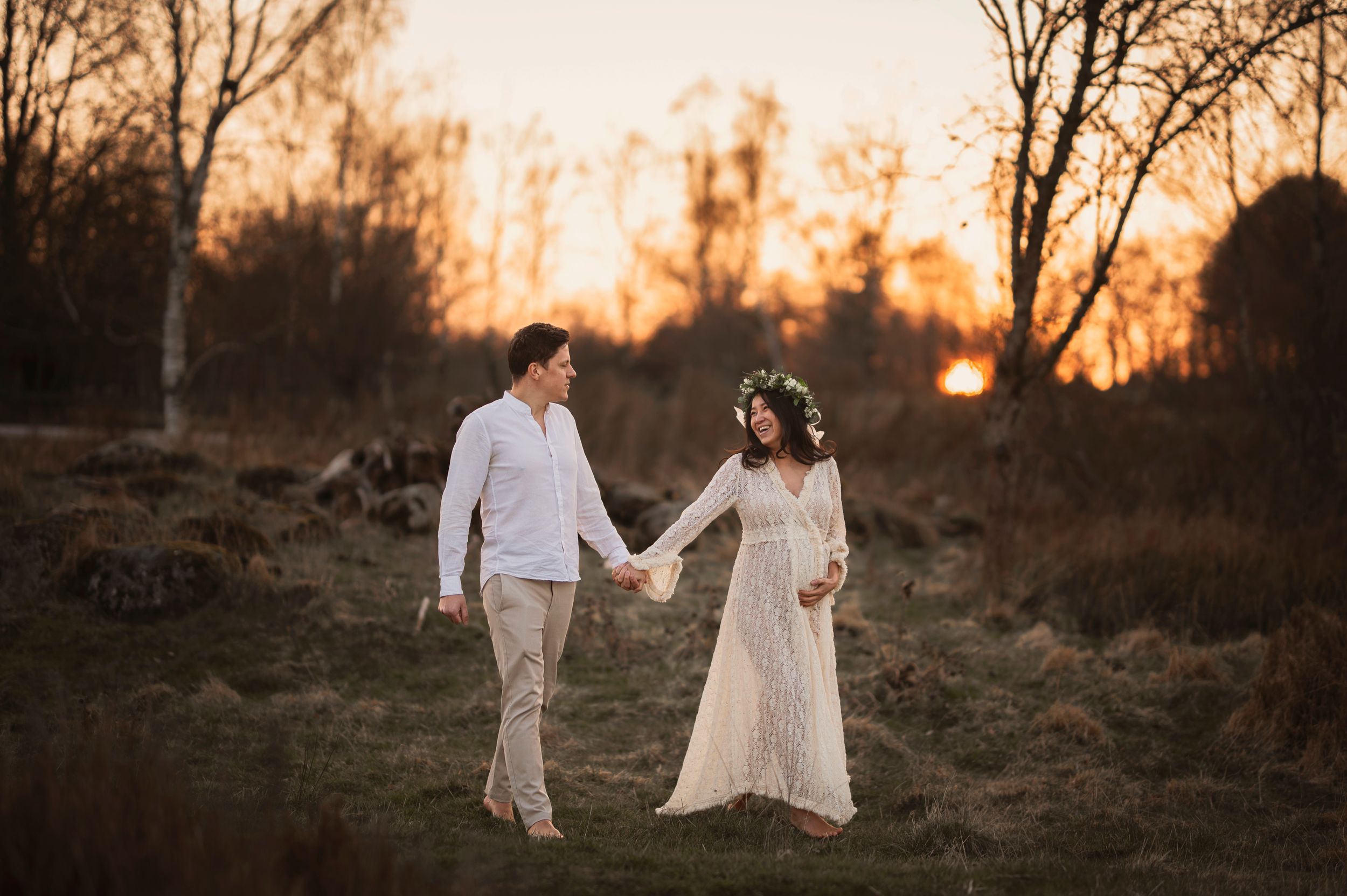 A couple in romantic white attire holds hands in a sunset-lit field surrounded by bare trees.