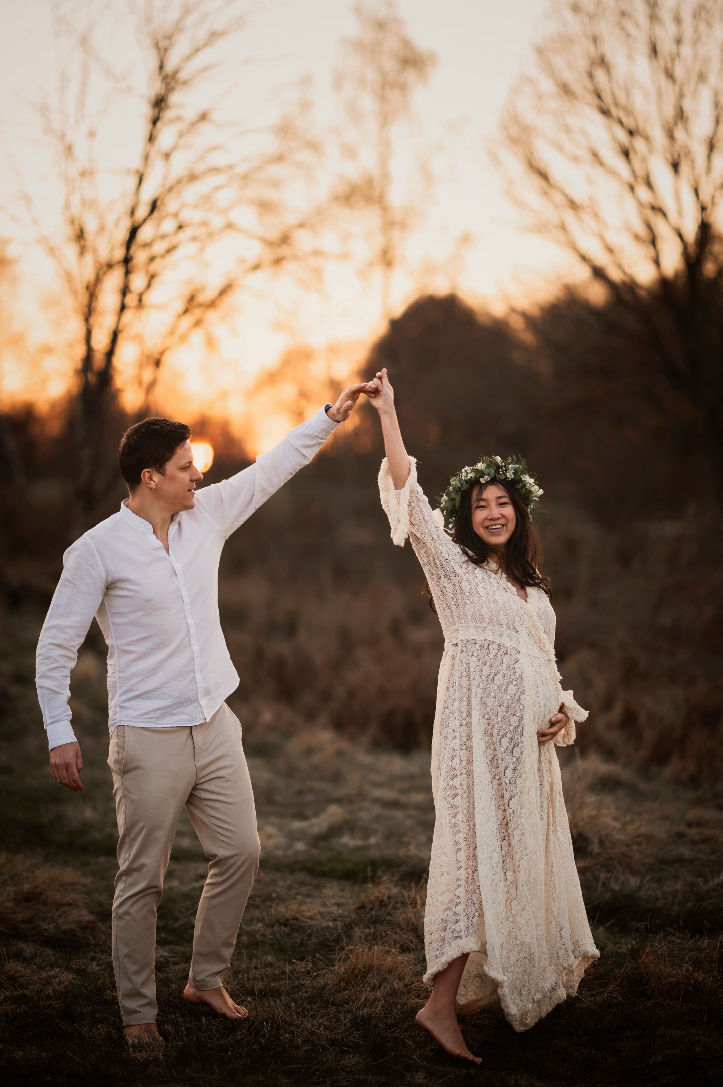 A couple dances barefoot in a field at sunset, with the woman wearing a flowing white lace dress and floral crown.