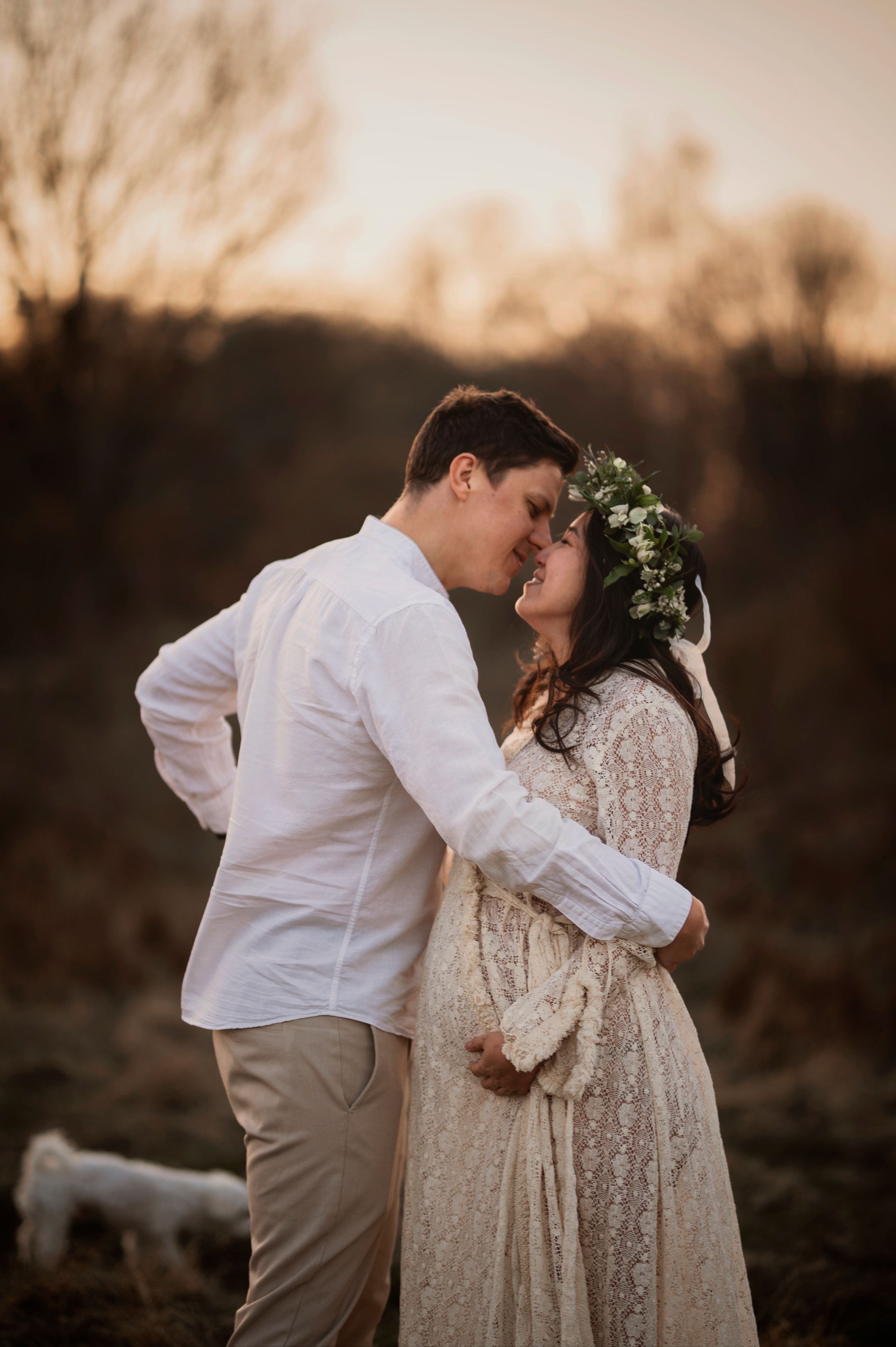A romantic sunset engagement photo of a couple in white clothing embracing outdoors with a floral crown.