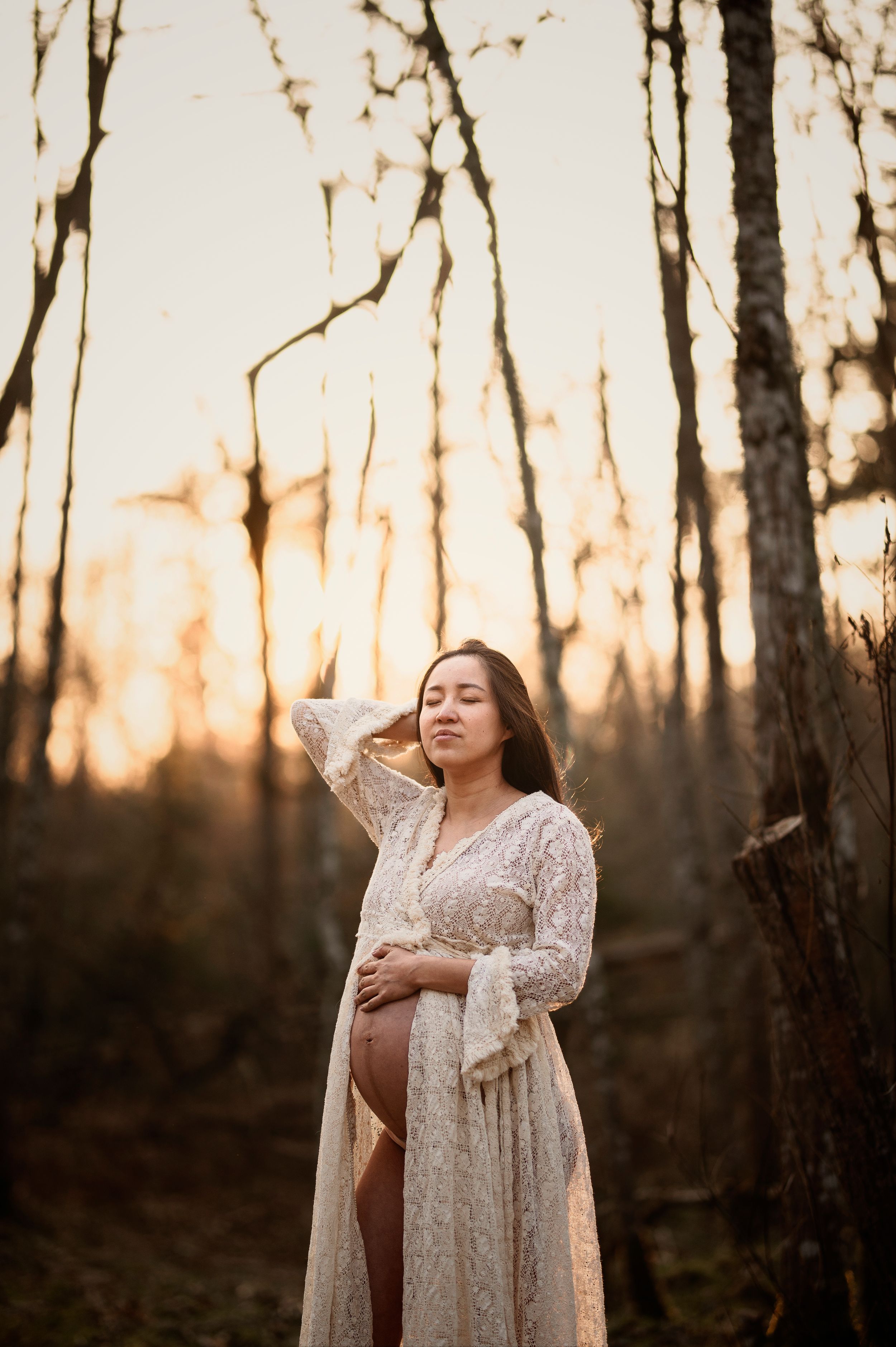A pregnant woman in a lace dress stands in a forest at sunset with warm golden light filtering through the trees.