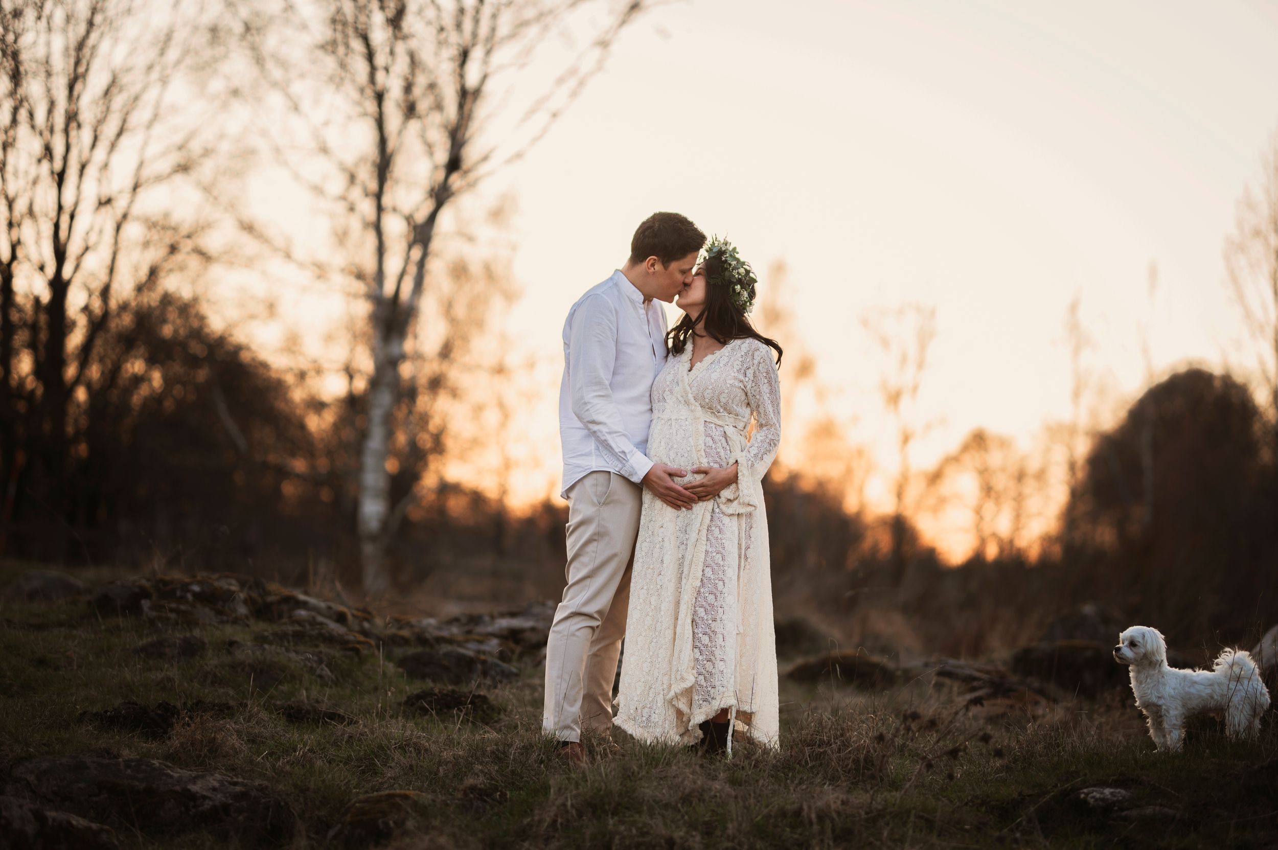 A couple embraces in a white wedding attire at sunset while a small white dog stands nearby on a grassy hillside.
