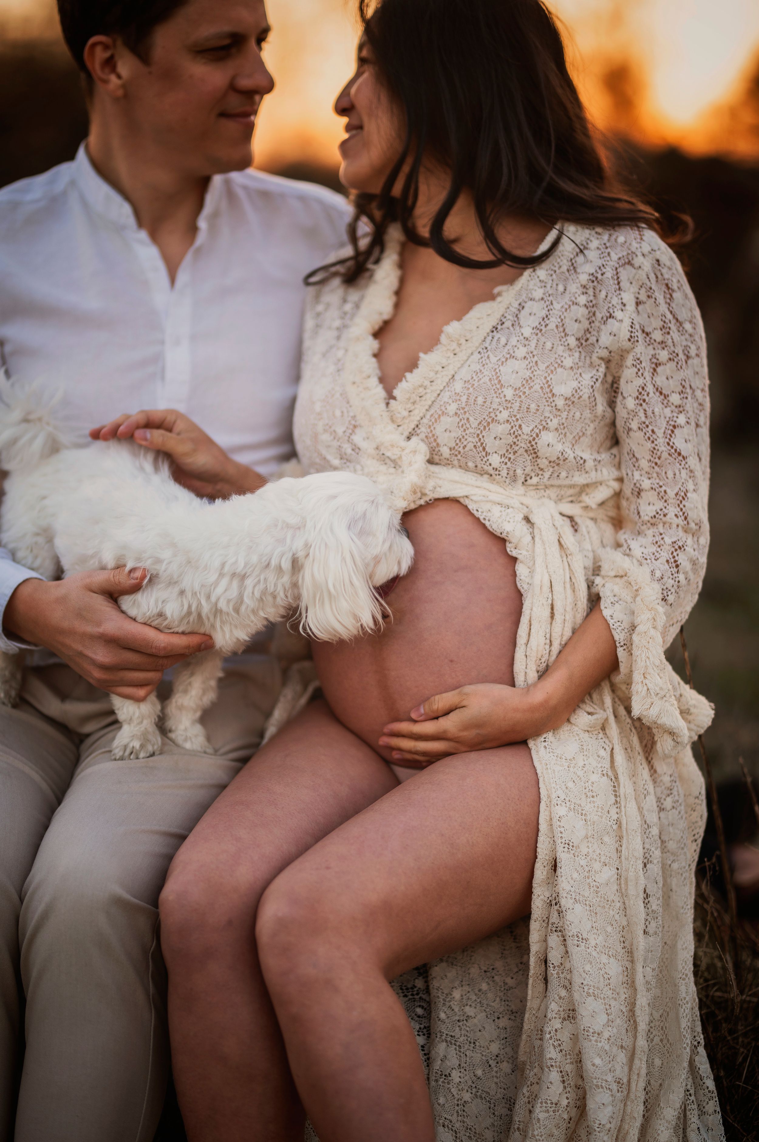 Intimate maternity photo during golden hour featuring a couple in white attire holding a small white fluffy dog.