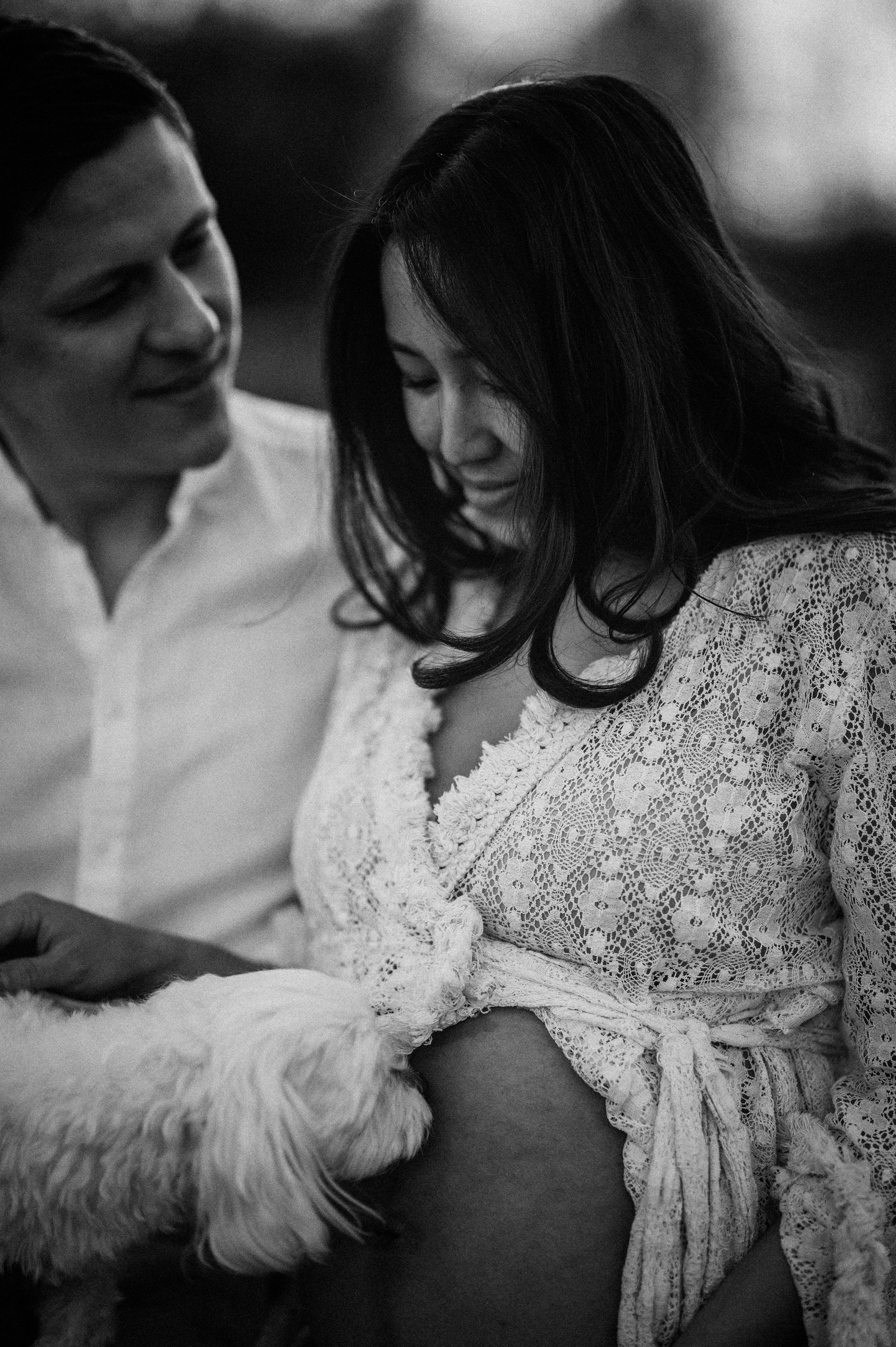 A tender black and white portrait of an expectant couple with a white dog sharing an intimate moment.