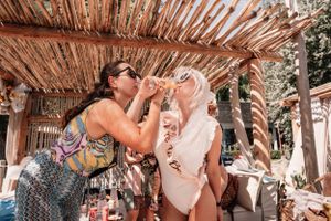 Two friends sharing drinks and laughing under rustic wooden beach bar covering.
