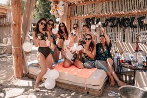 Group of friends in swimwear celebrating at beach club lounge area with balloons and drinks.