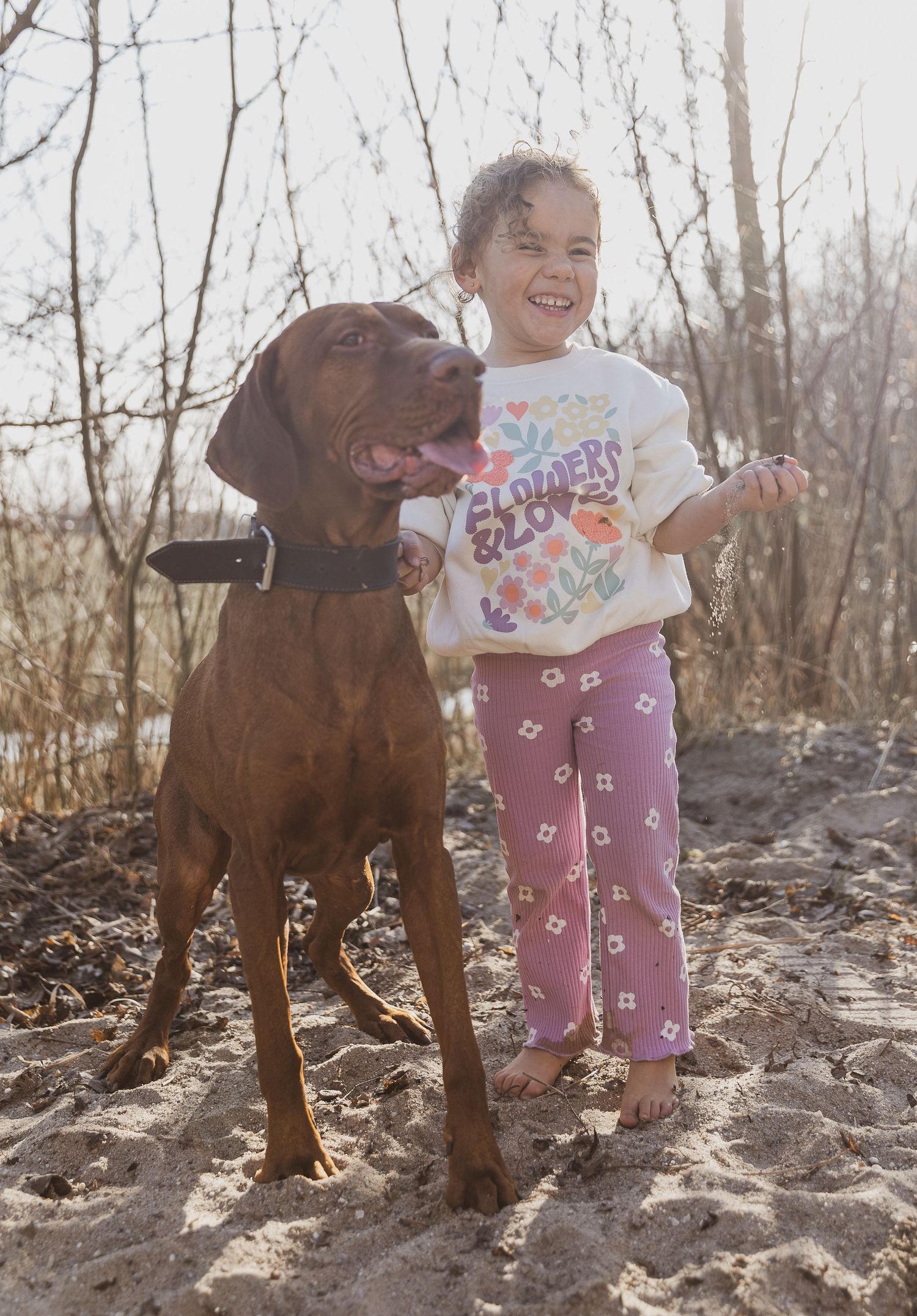 Child in colorful shirt stands joyfully with large brown dog in natural setting. kinderportretten fotograaf