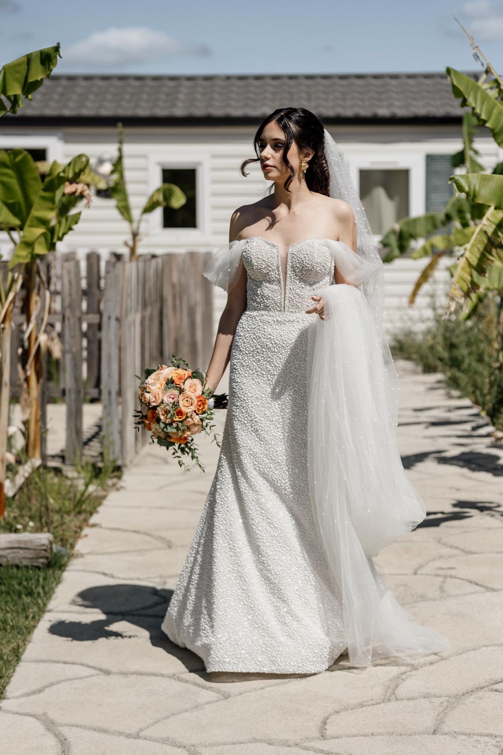 Beautiful wedding dress with off-shoulder neckline and lace details, accessorized with a flowing veil and peach-toned bouquet.