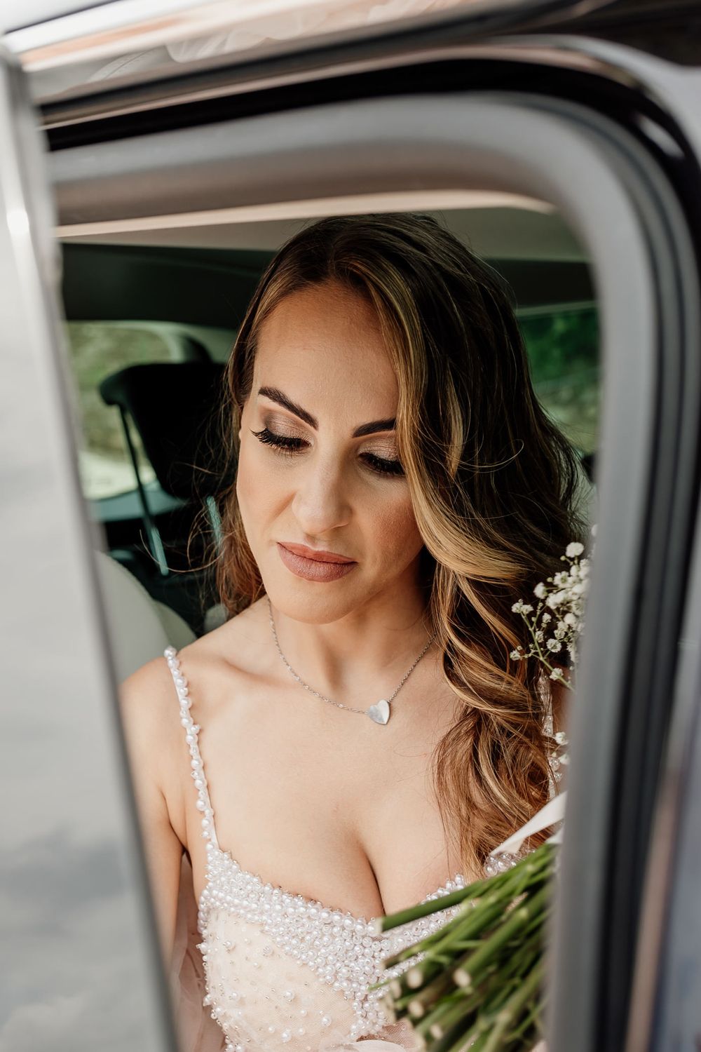 A bride wearing a sparkly white wedding dress and delicate hair accessories looks down thoughtfully through a car window.