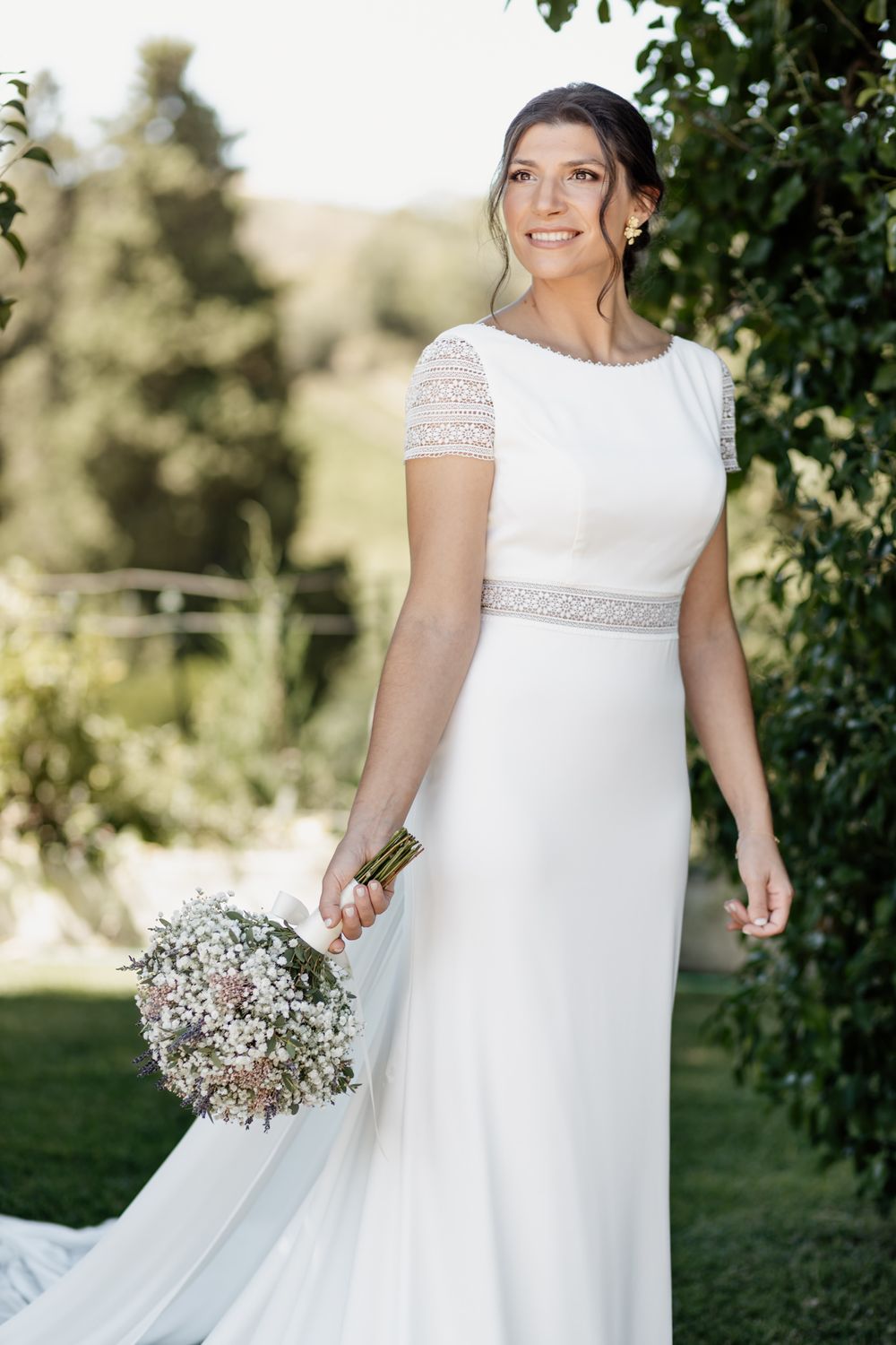 A bride in an elegant white wedding dress with lace details holds a baby's breath bouquet in a garden setting.