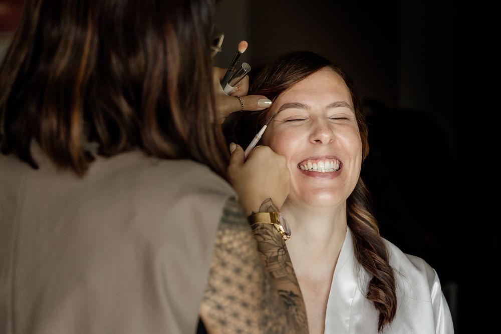 Makeup artist applies cosmetics on smiling client wearing white blouse during beauty session.