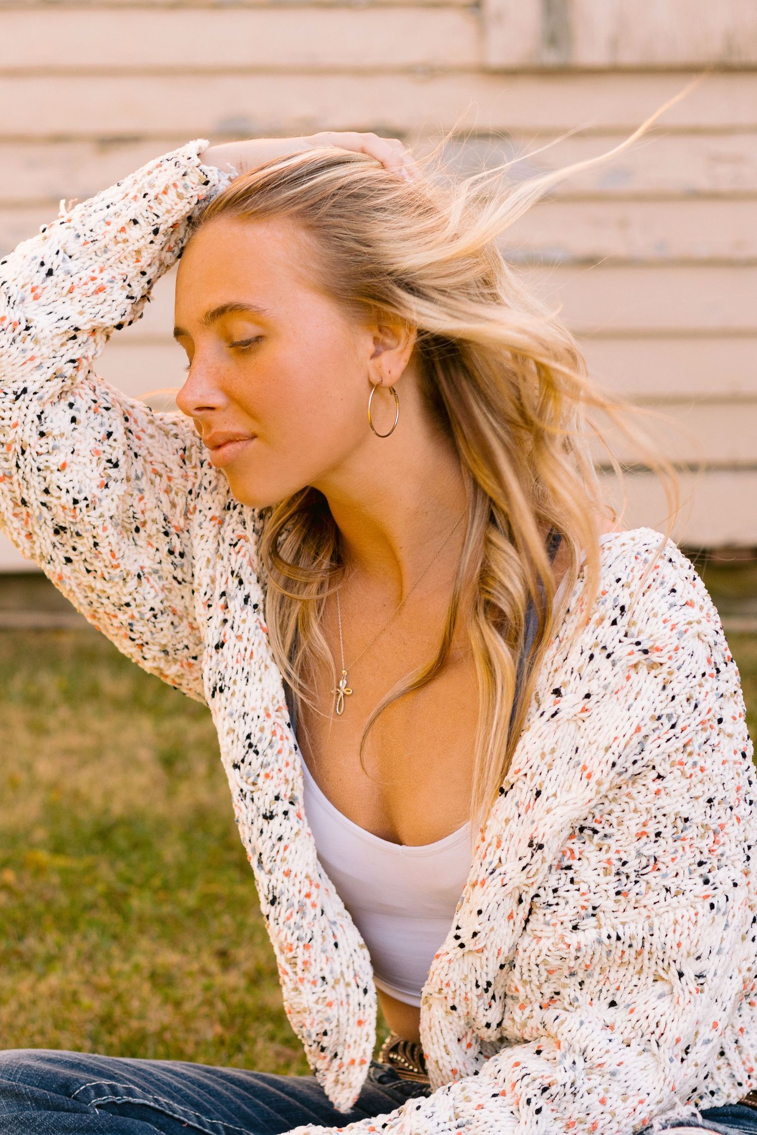 Someone wearing a speckled white knit cardigan and tank top poses outdoors with windblown blonde hair.