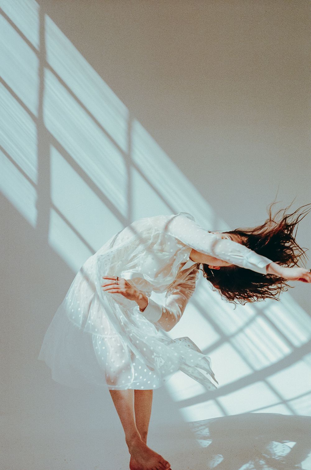 Figure in white dress bends dramatically in shaft of window light creating geometric shadows.