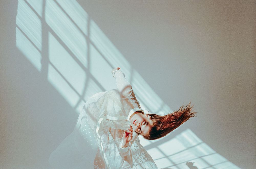 A figure in white clothing lies gracefully against a wall with dramatic window light patterns casting shadows.