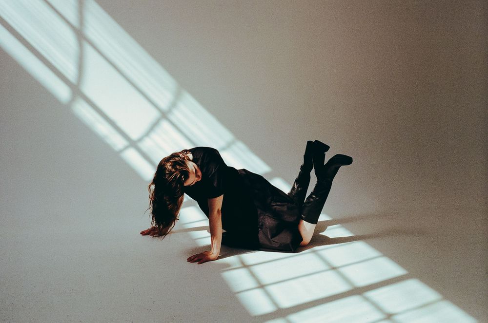Figure in black clothing poses dramatically on floor with geometric window shadows.