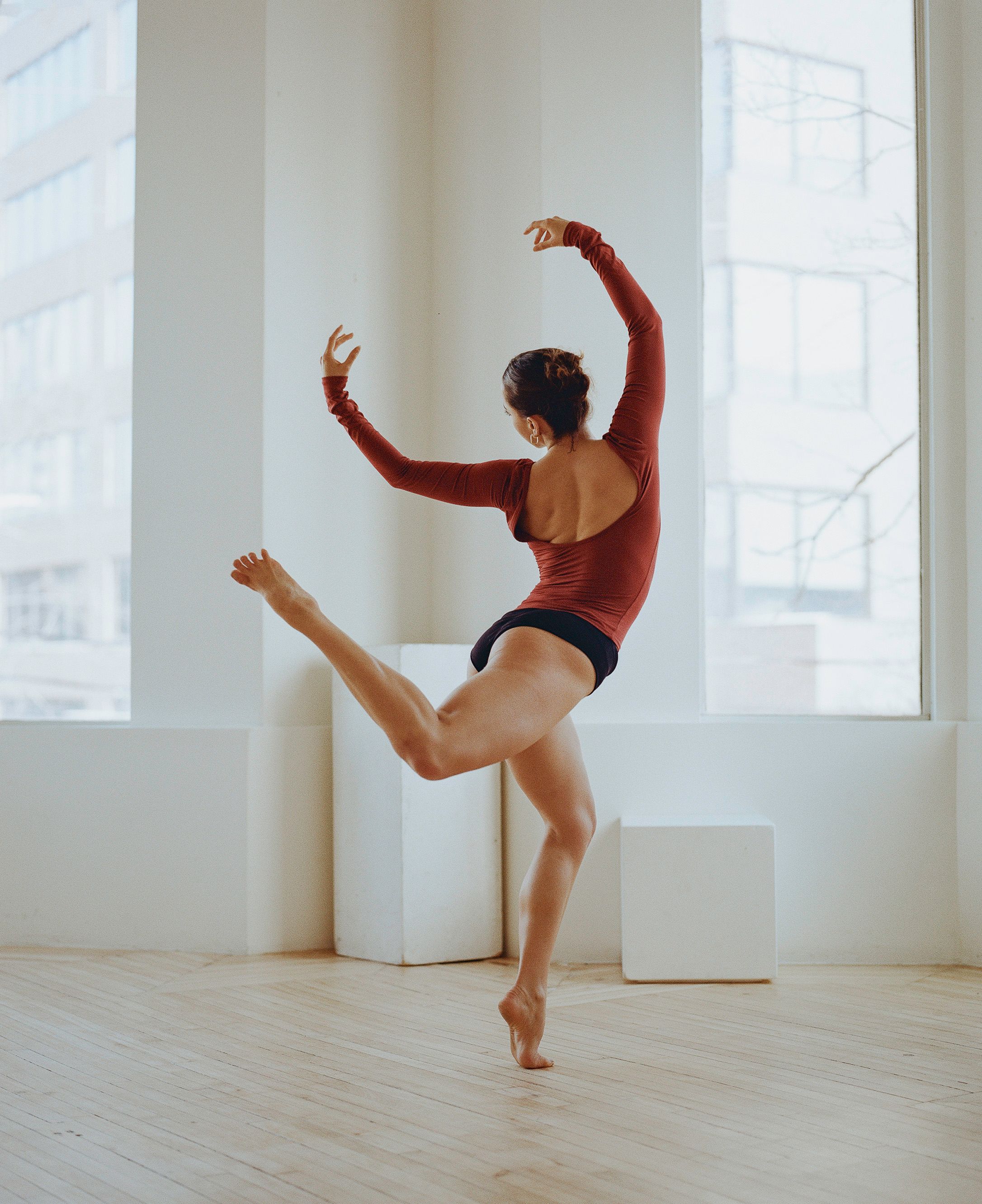 Modern dancer in red leotard performs graceful movement in sunlit studio space.