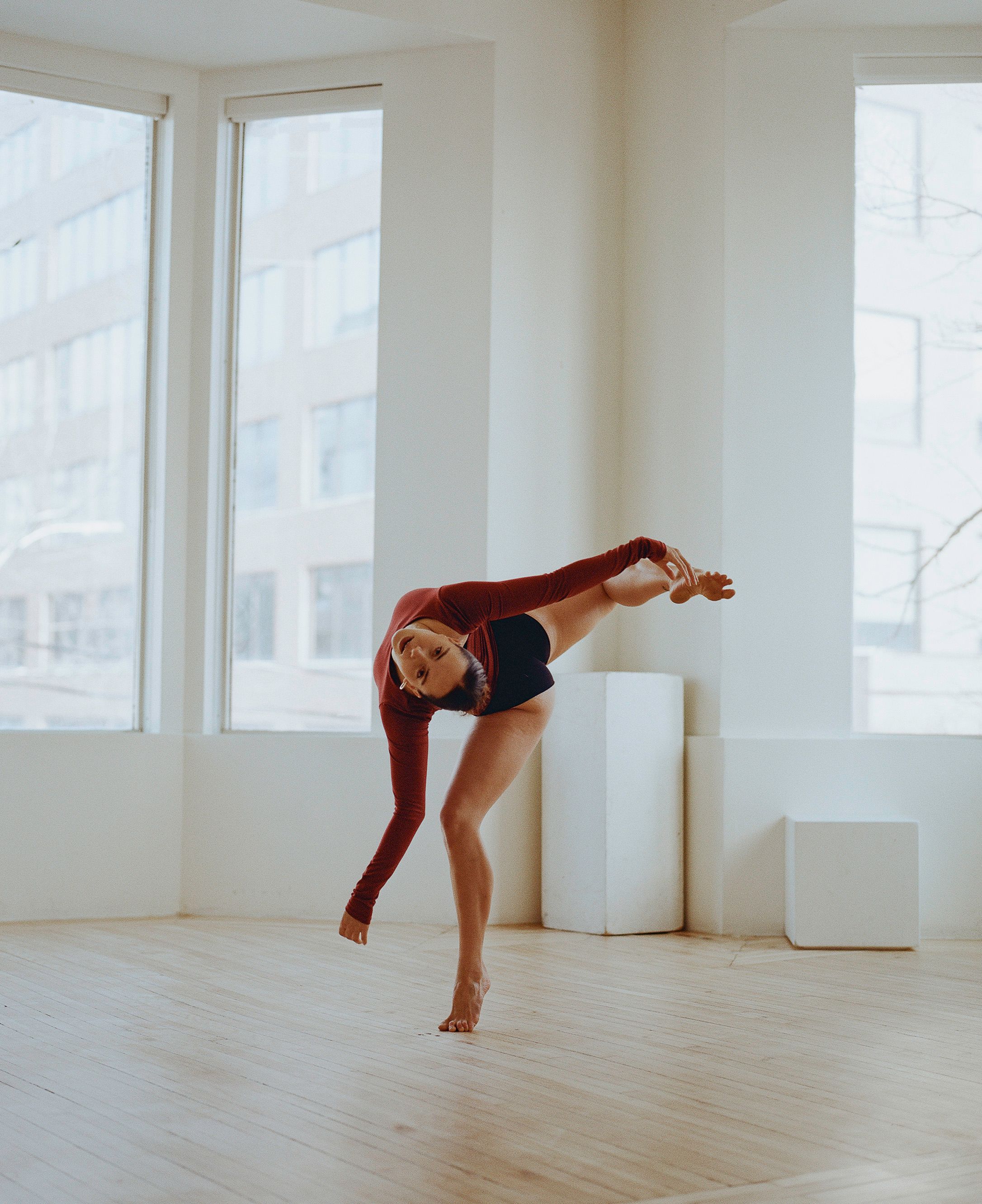 Dancer in rust-colored leotard performs expressive movement in bright studio space.