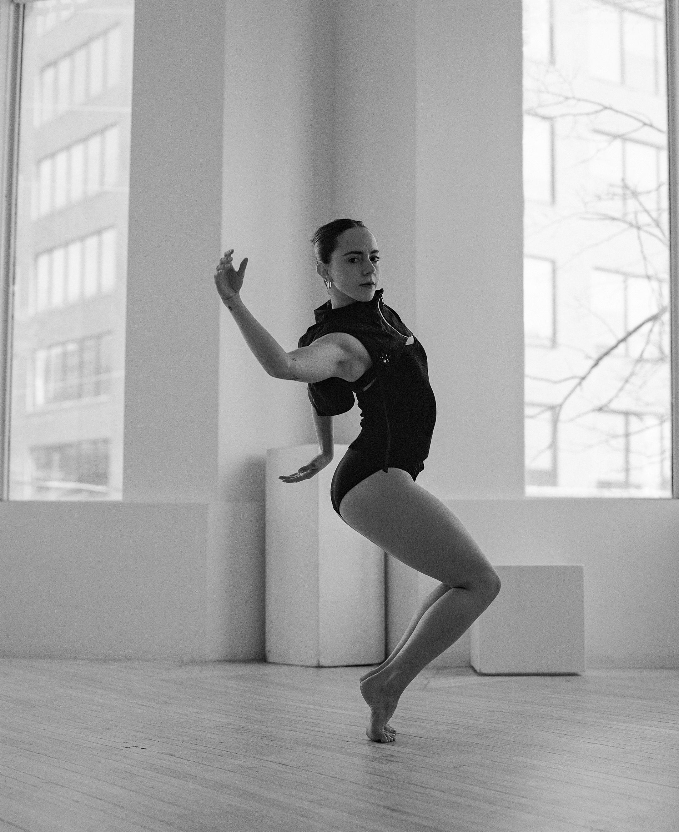 Modern dancer strikes dynamic pose against bright windows in minimalist studio setting.