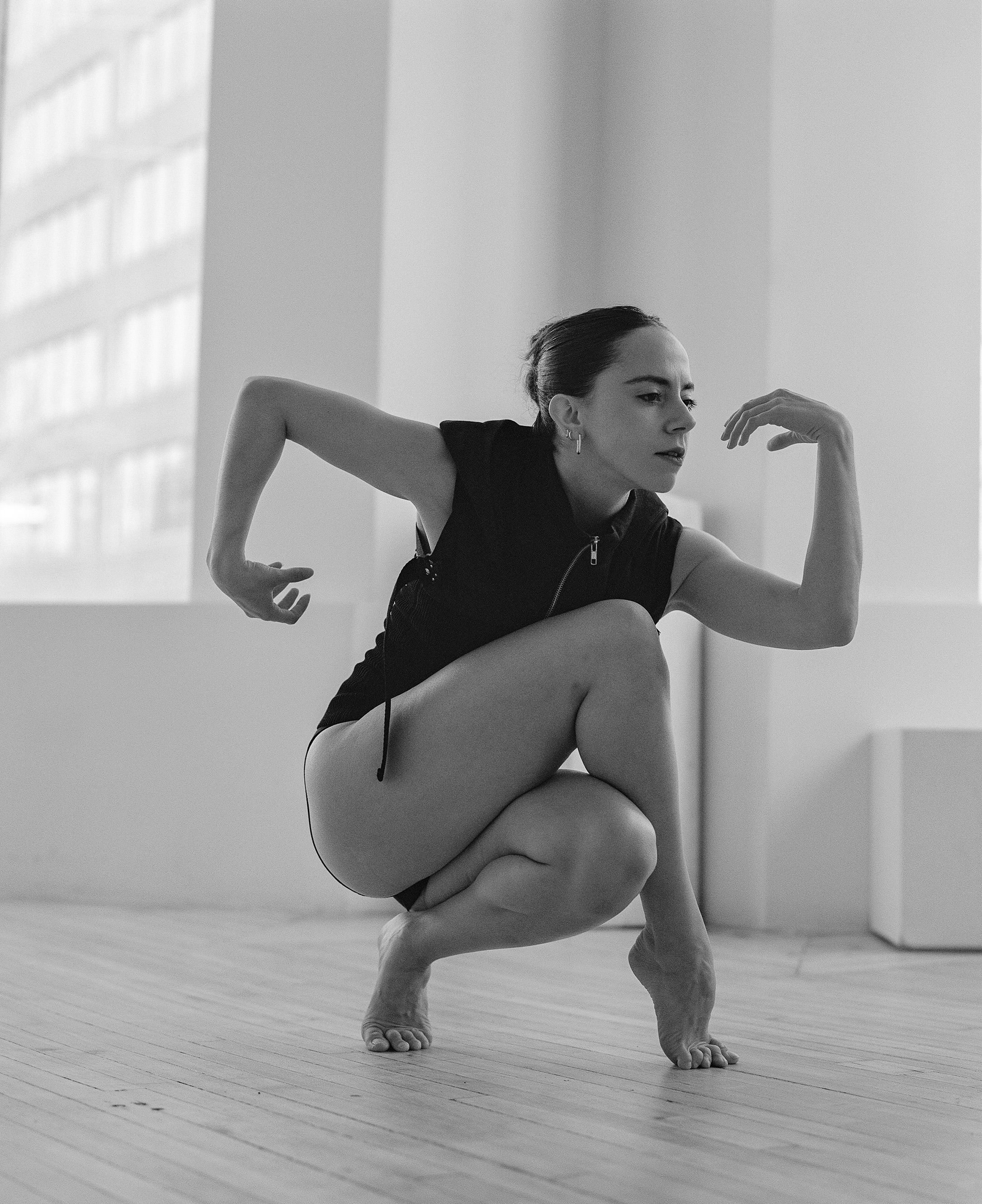 Contemporary dancer in black leotard performs dynamic squatting pose on wooden floor in bright studio.