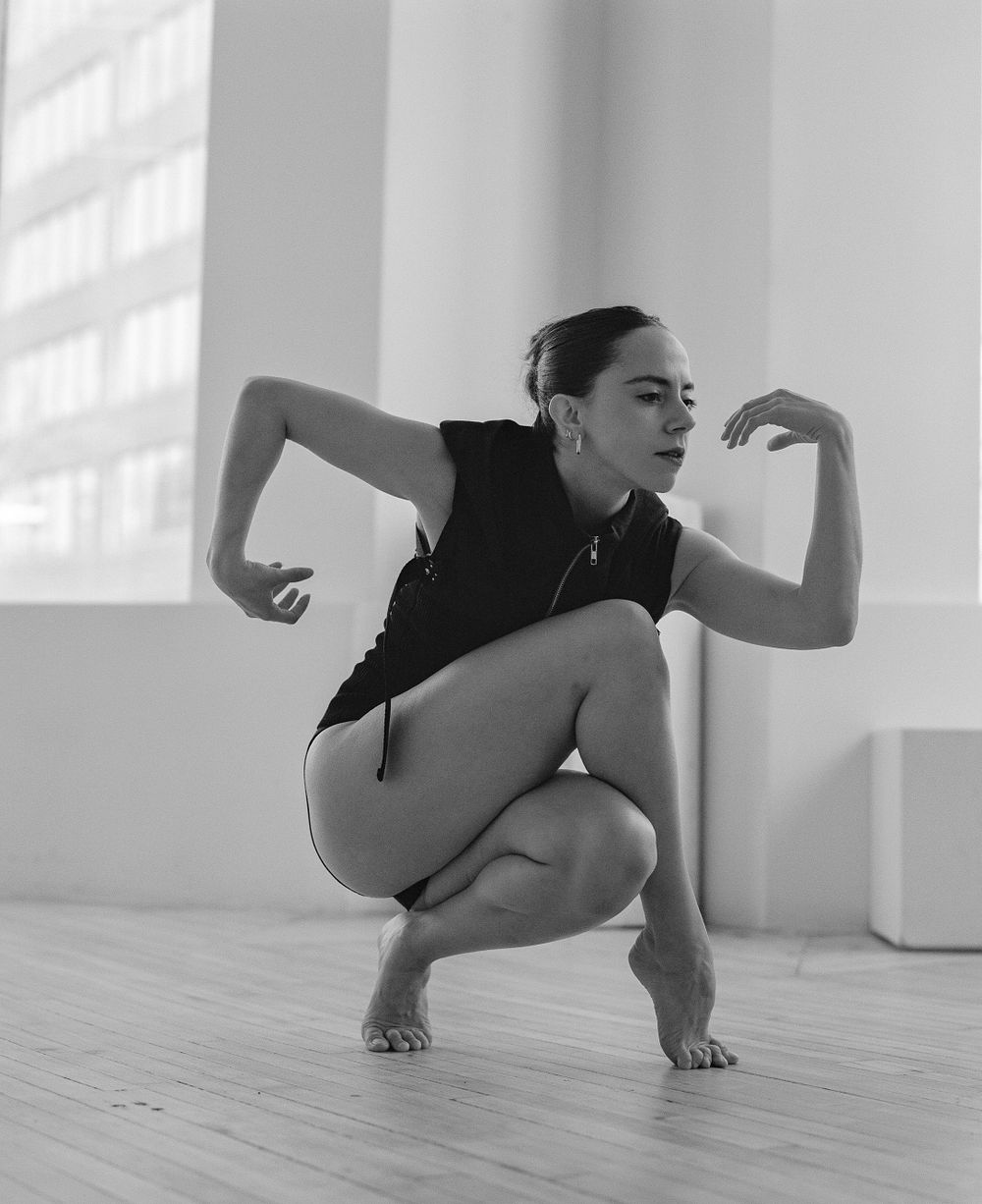 Contemporary dancer in black leotard performs dynamic squatting pose on wooden floor in bright studio.