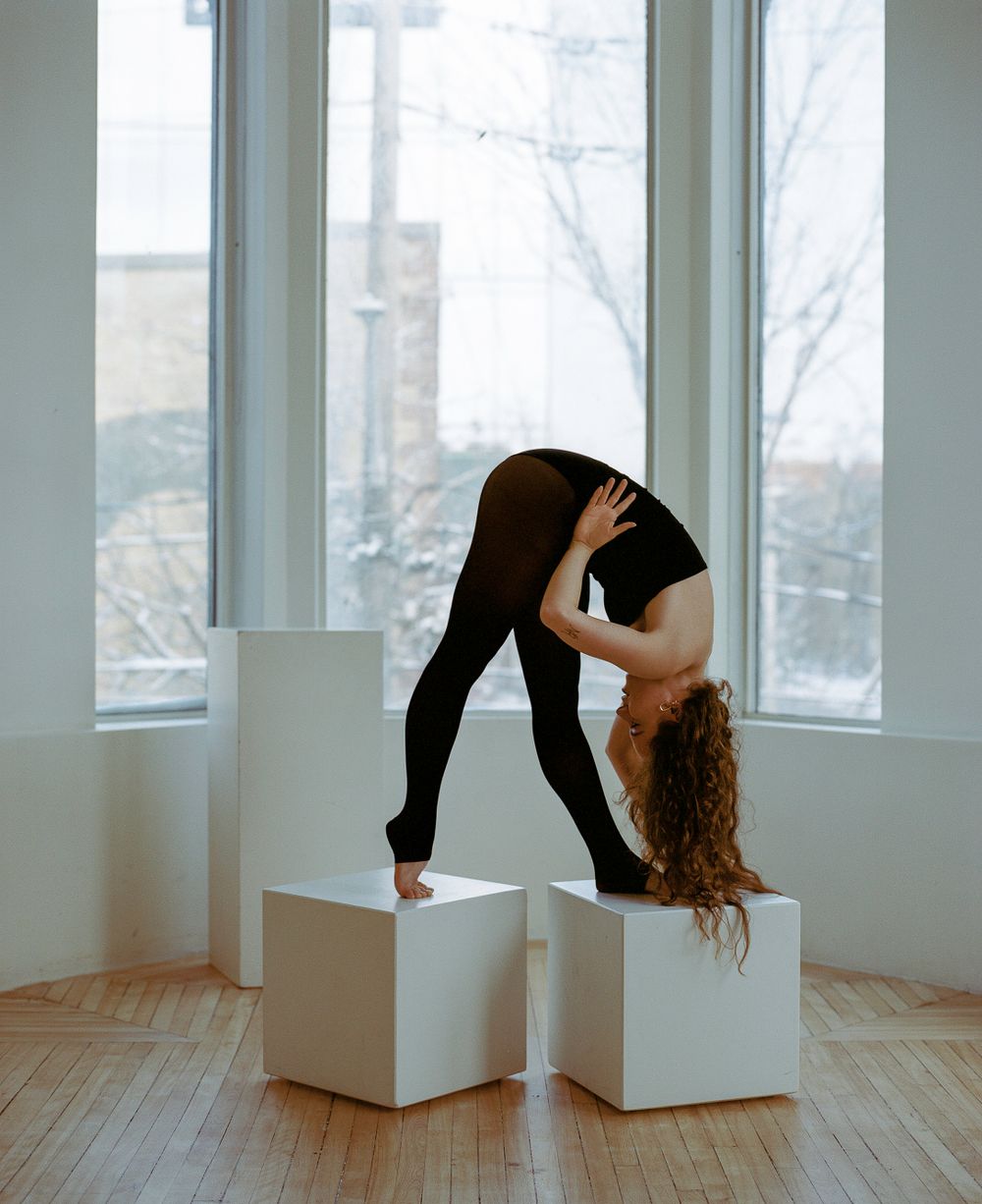 Modern dancer performs split pose across two white cubes near sunlit windows.